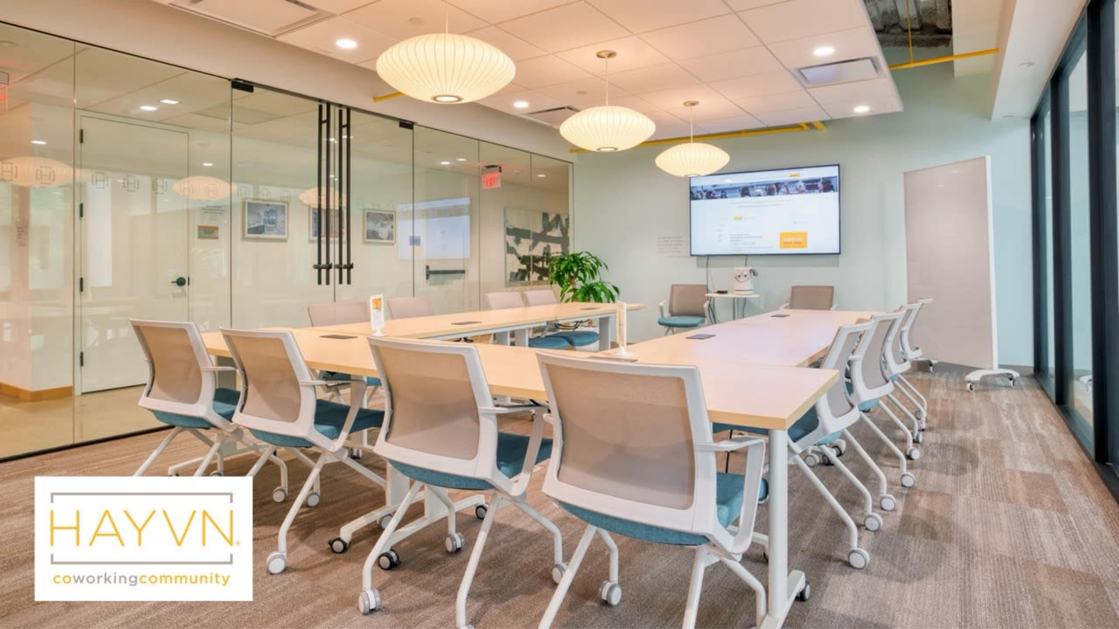 A modern conference room features a long table with ergonomic chairs, surrounded by glass walls and a large screen for presentations.