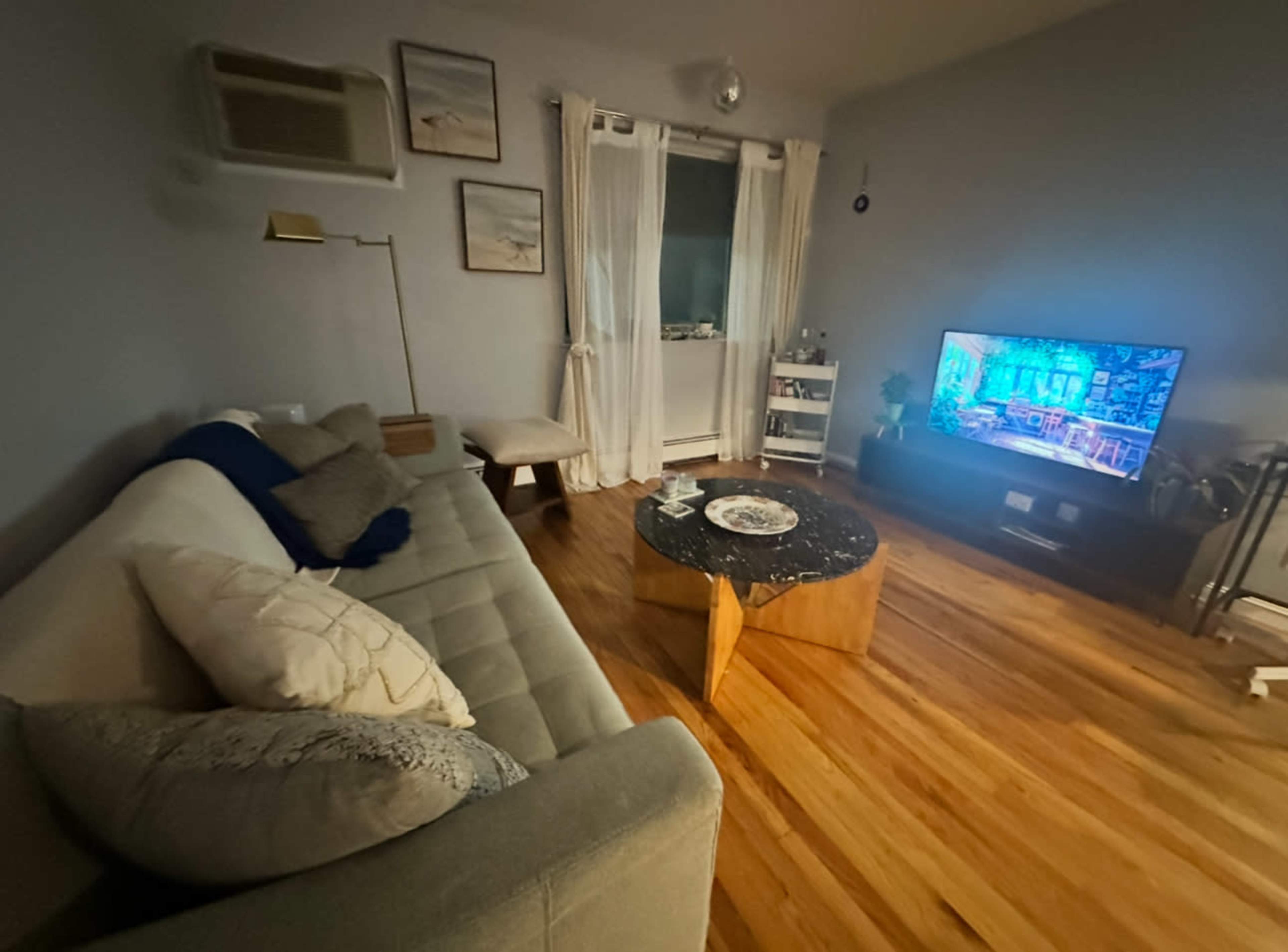 The image shows a living room featuring a sofa, a coffee table, a television, and a window with sheer curtains.