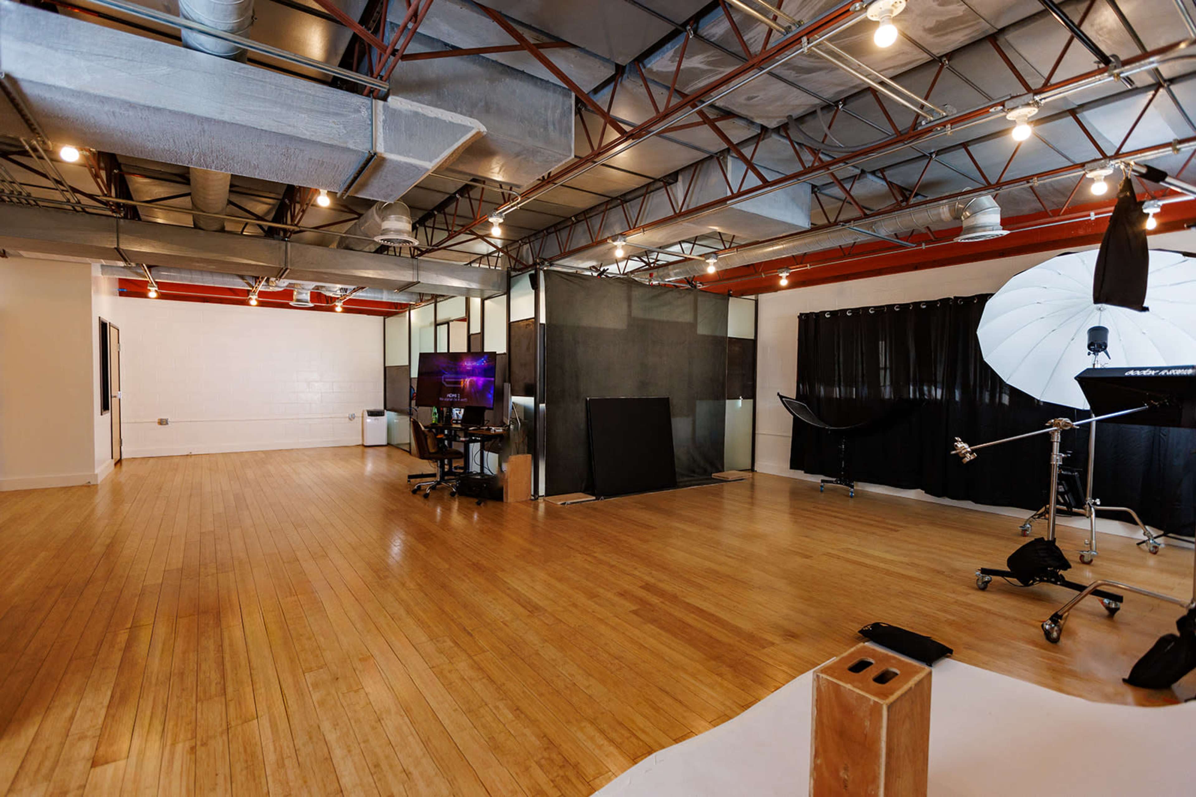 The image shows a spacious studio with wooden flooring, large windows, and a variety of equipment set up for photography.