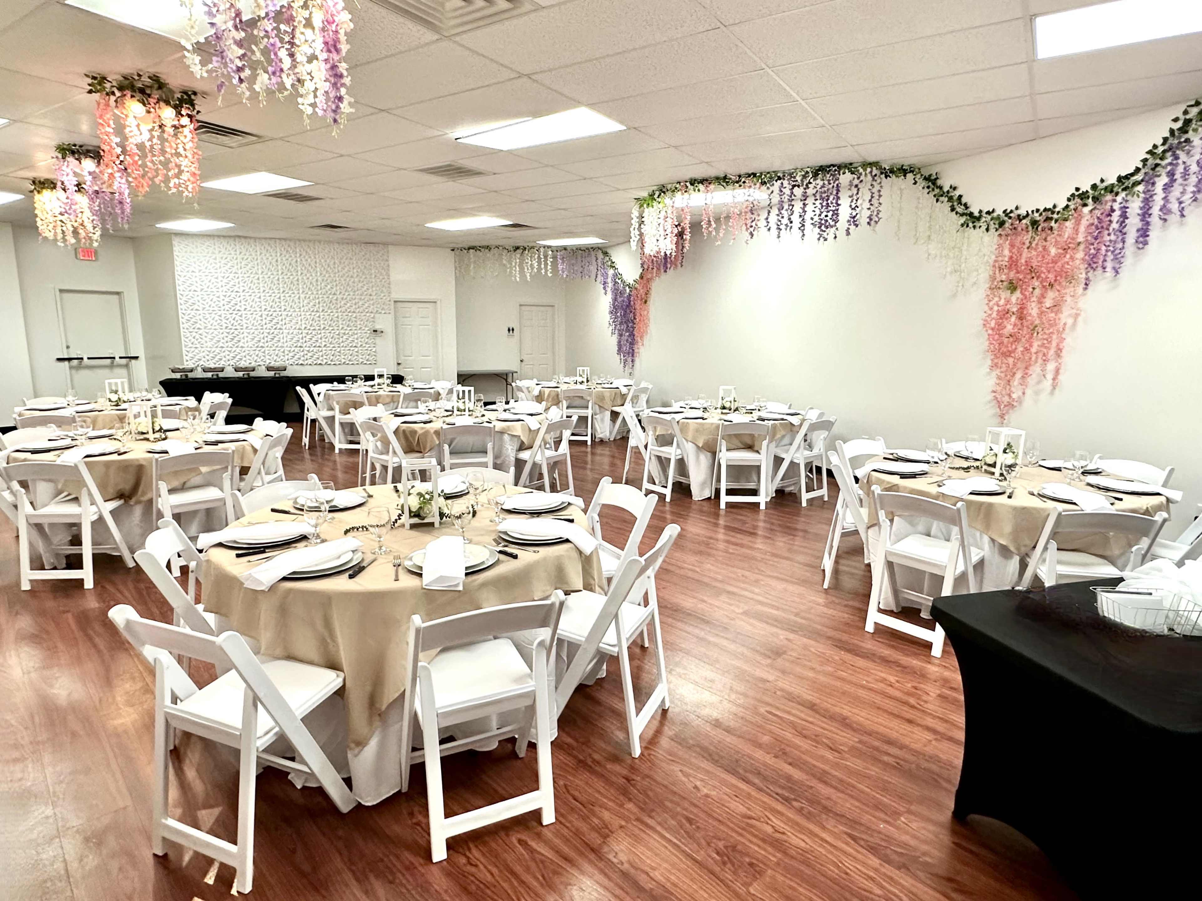 A banquet hall arranged with round tables covered in beige tablecloths, set for dining, and adorned with floral decorations hanging from the ceiling.