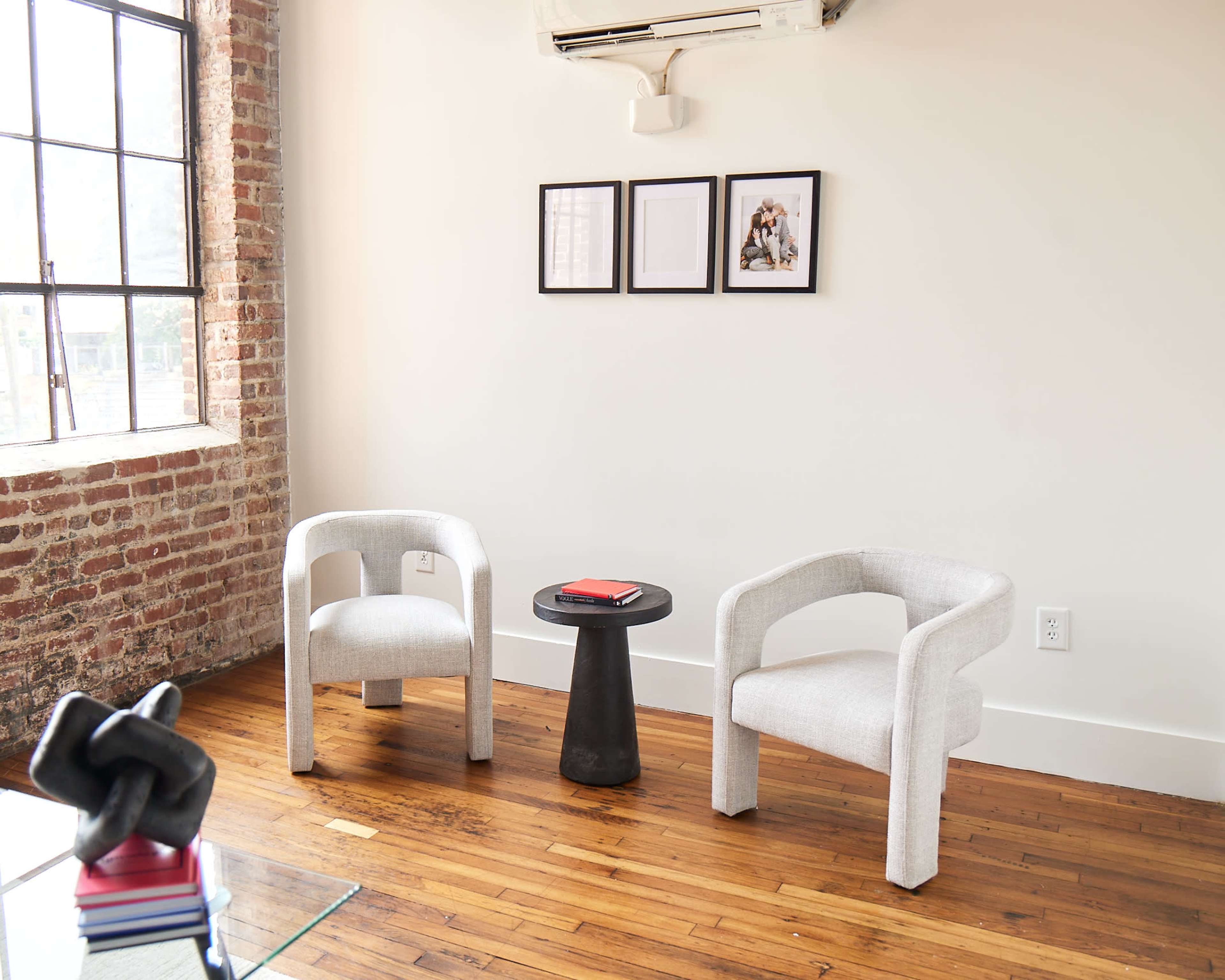 A minimalistic interior space featuring two light-colored chairs, a small black side table, and three framed pictures on the wall beside a large window.