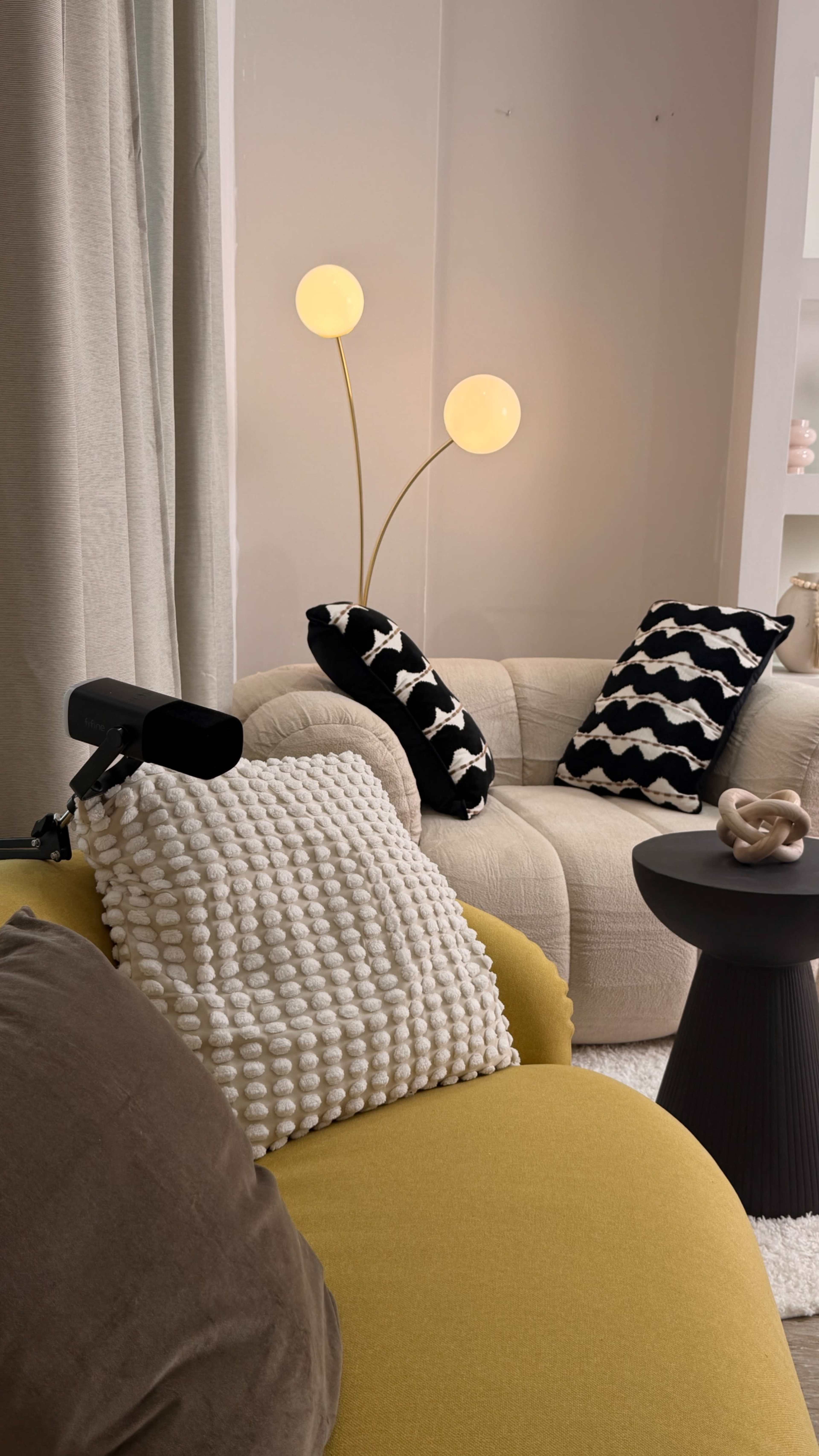 The image shows a cozy living room scene featuring a yellow chair, a white couch with patterned cushions, and a black side table with a small decorative item.