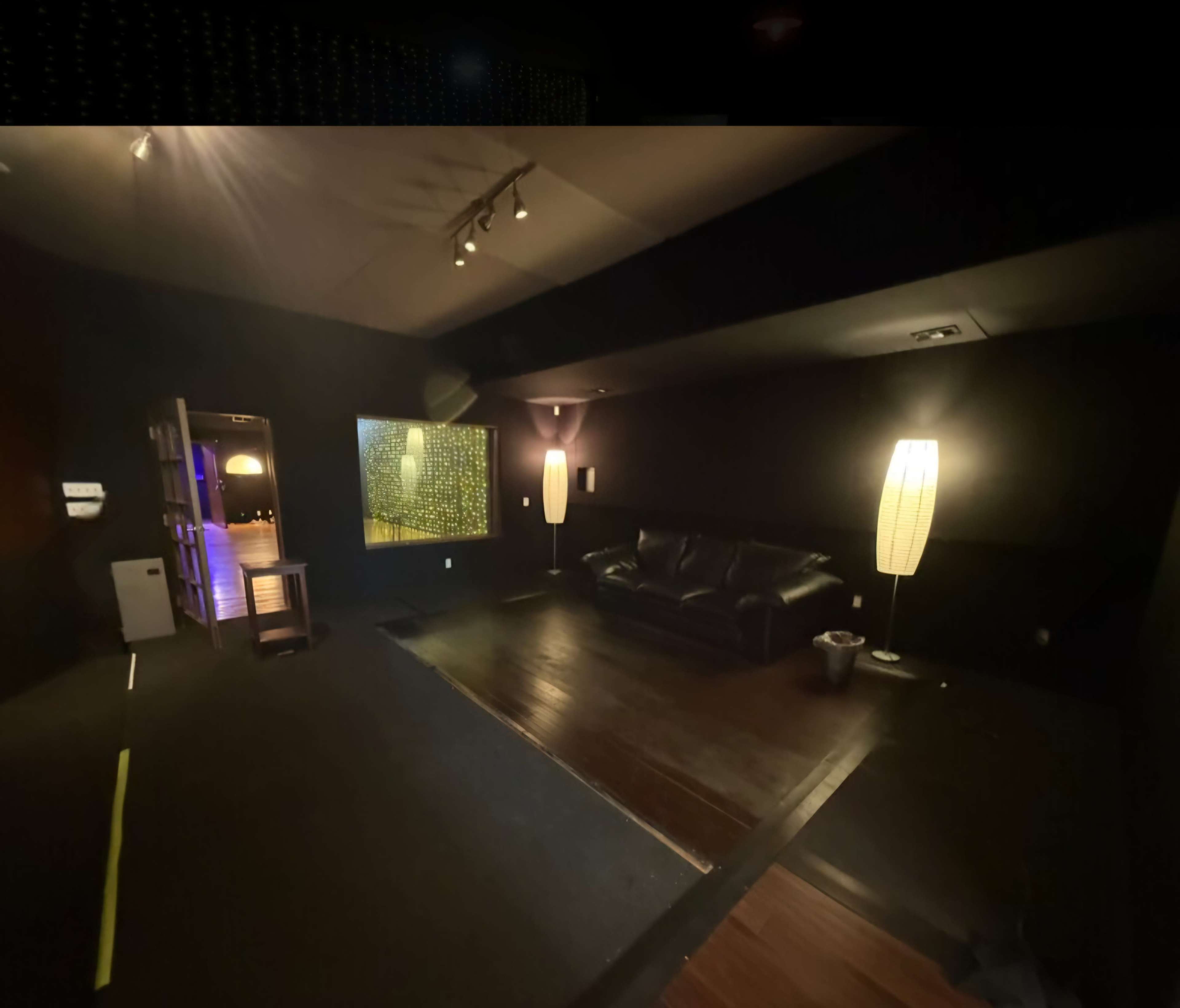 The image shows a dimly lit room with a black color scheme, featuring a large couch, a floor lamp, and a window displaying a lit interior in the background.