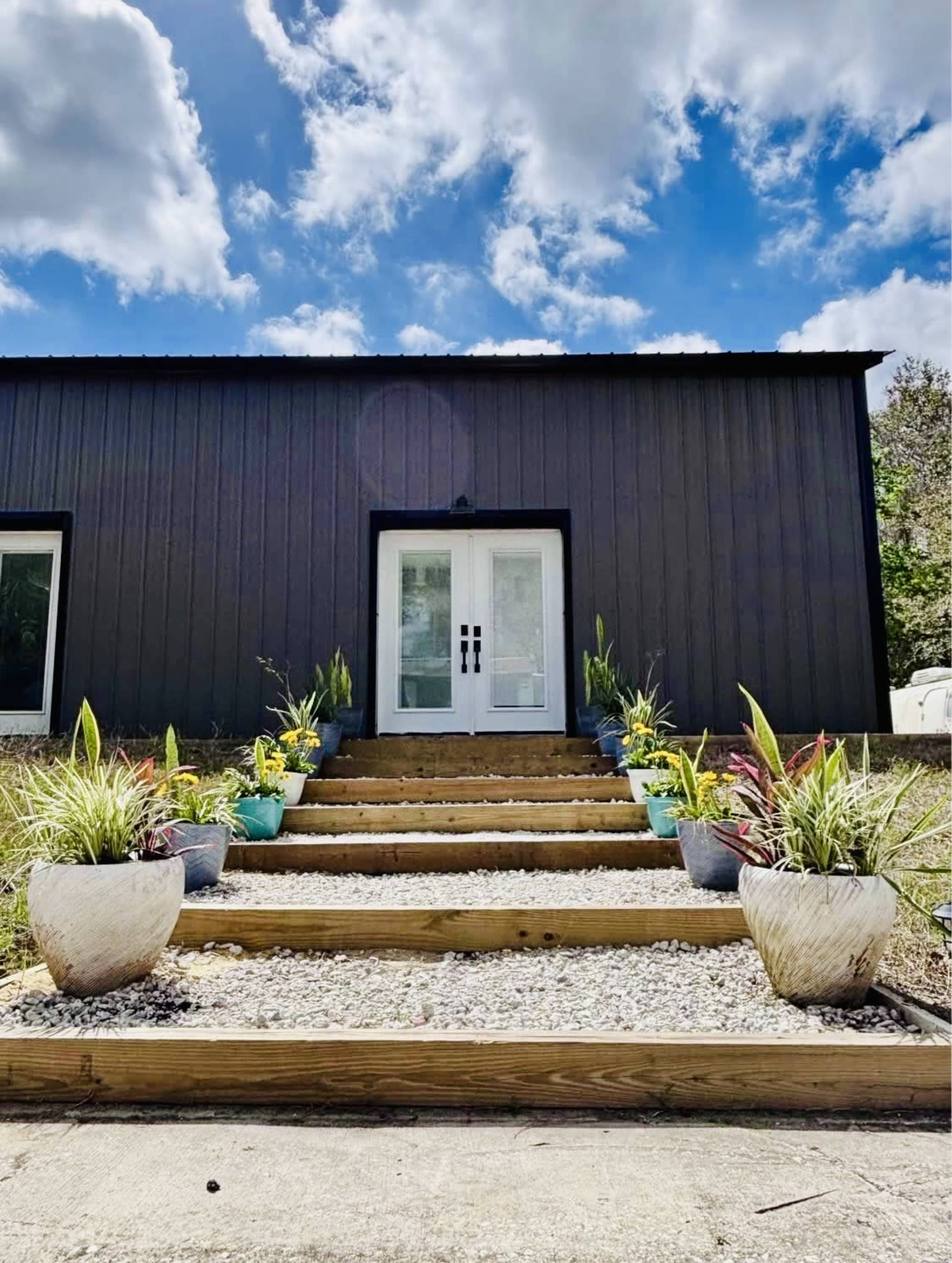 A set of wooden steps leads to a modern black building with double doors, flanked by various potted plants under a partly cloudy sky.