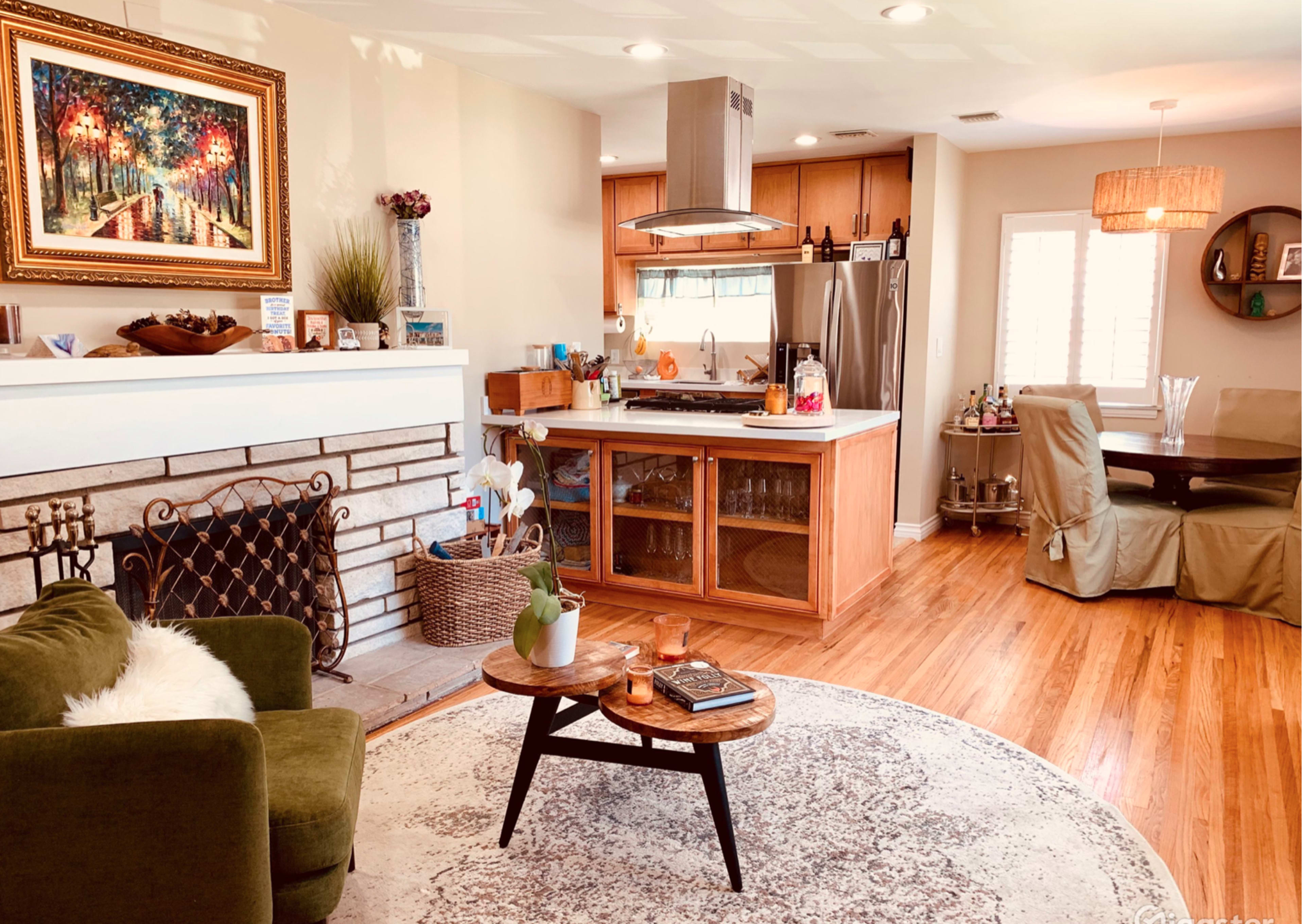 The image shows a cozy living room with a green sofa, a round coffee table, and a kitchen area in the background featuring wooden cabinetry and stainless steel appliances.