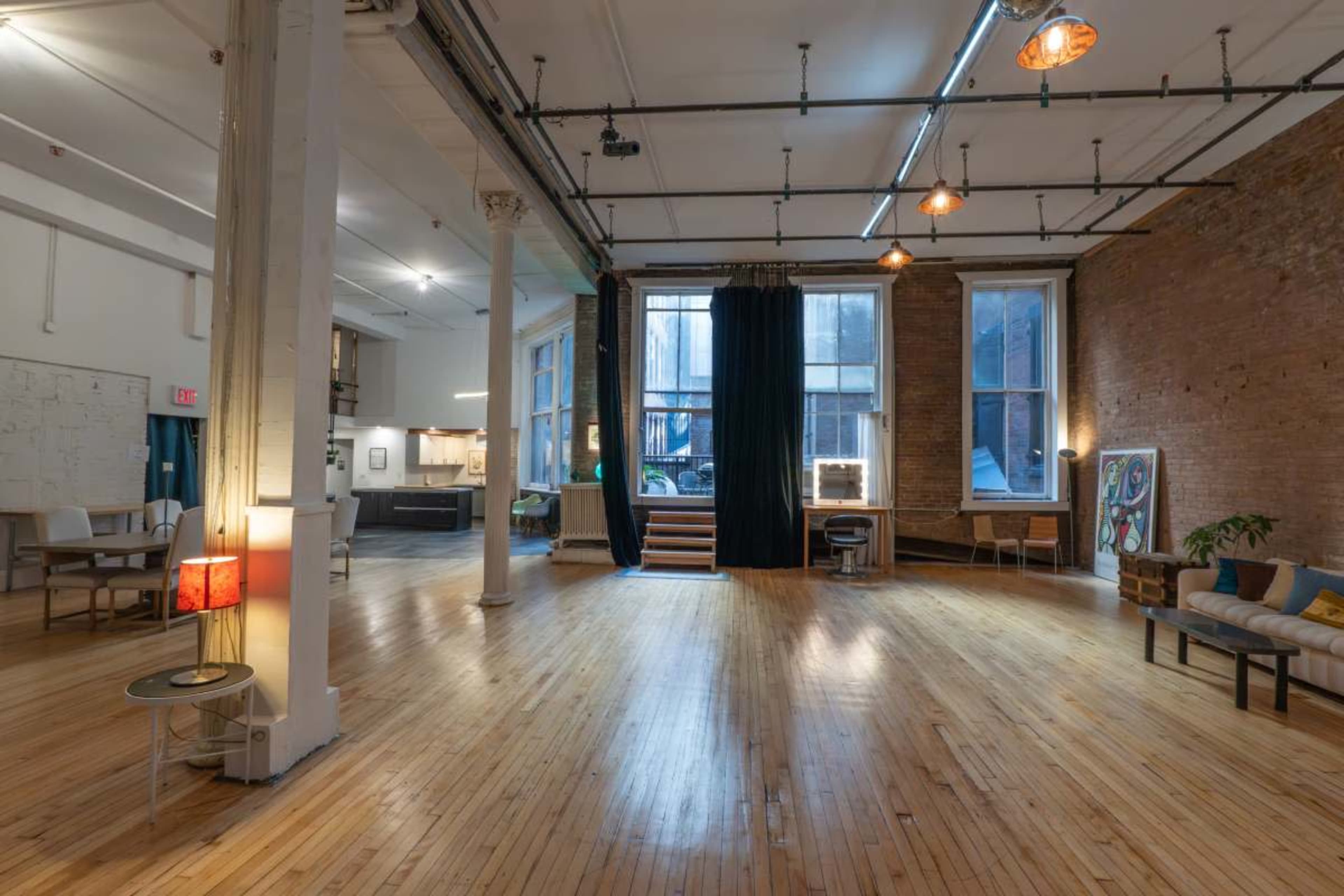 The image shows a spacious loft with wooden floors and large windows, featuring minimalistic furniture and a black curtain in the corner.