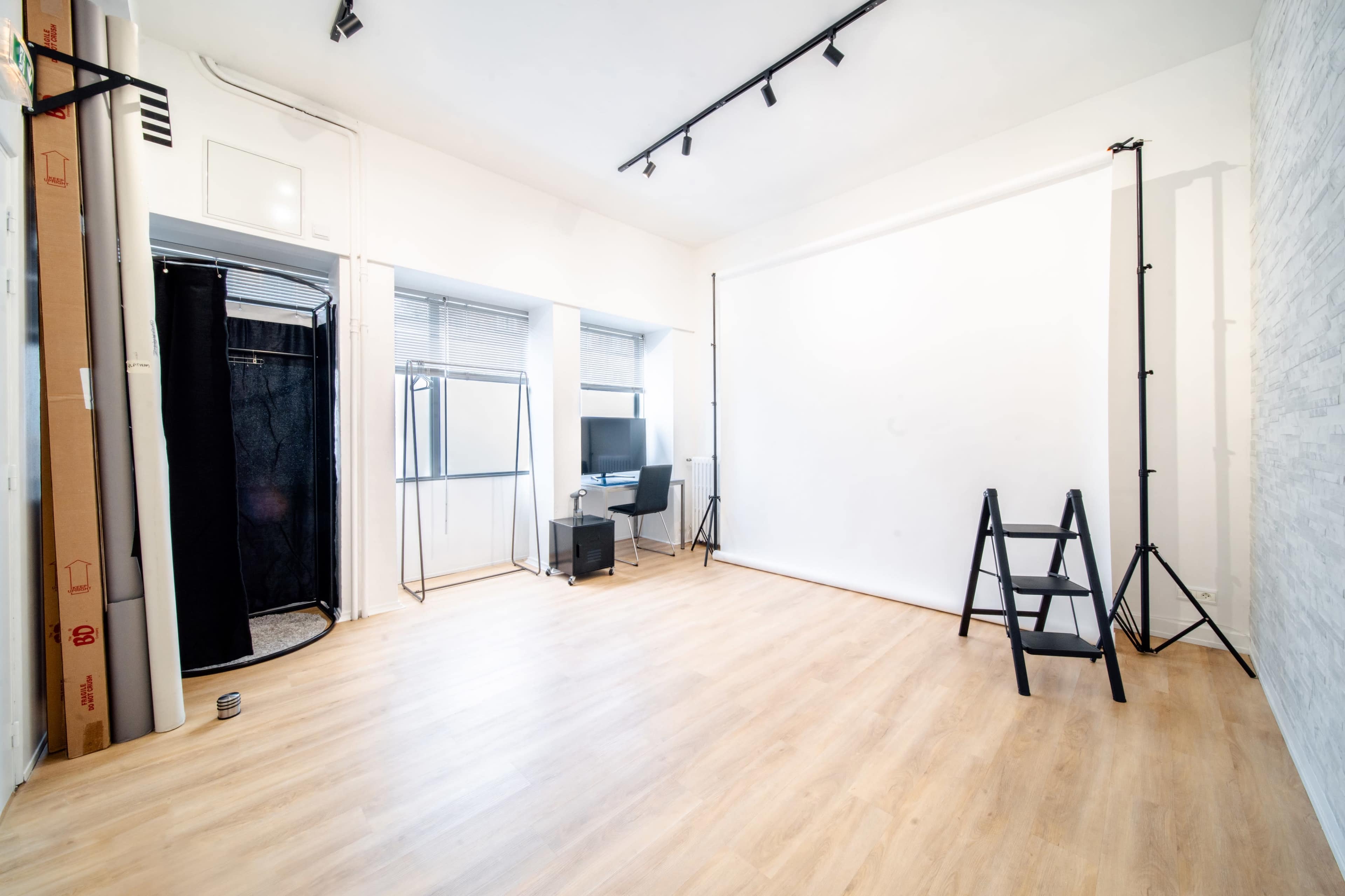 The image shows a spacious, well-lit photography studio with wooden flooring, a blank white backdrop, lighting equipment, and a television on a black stand.