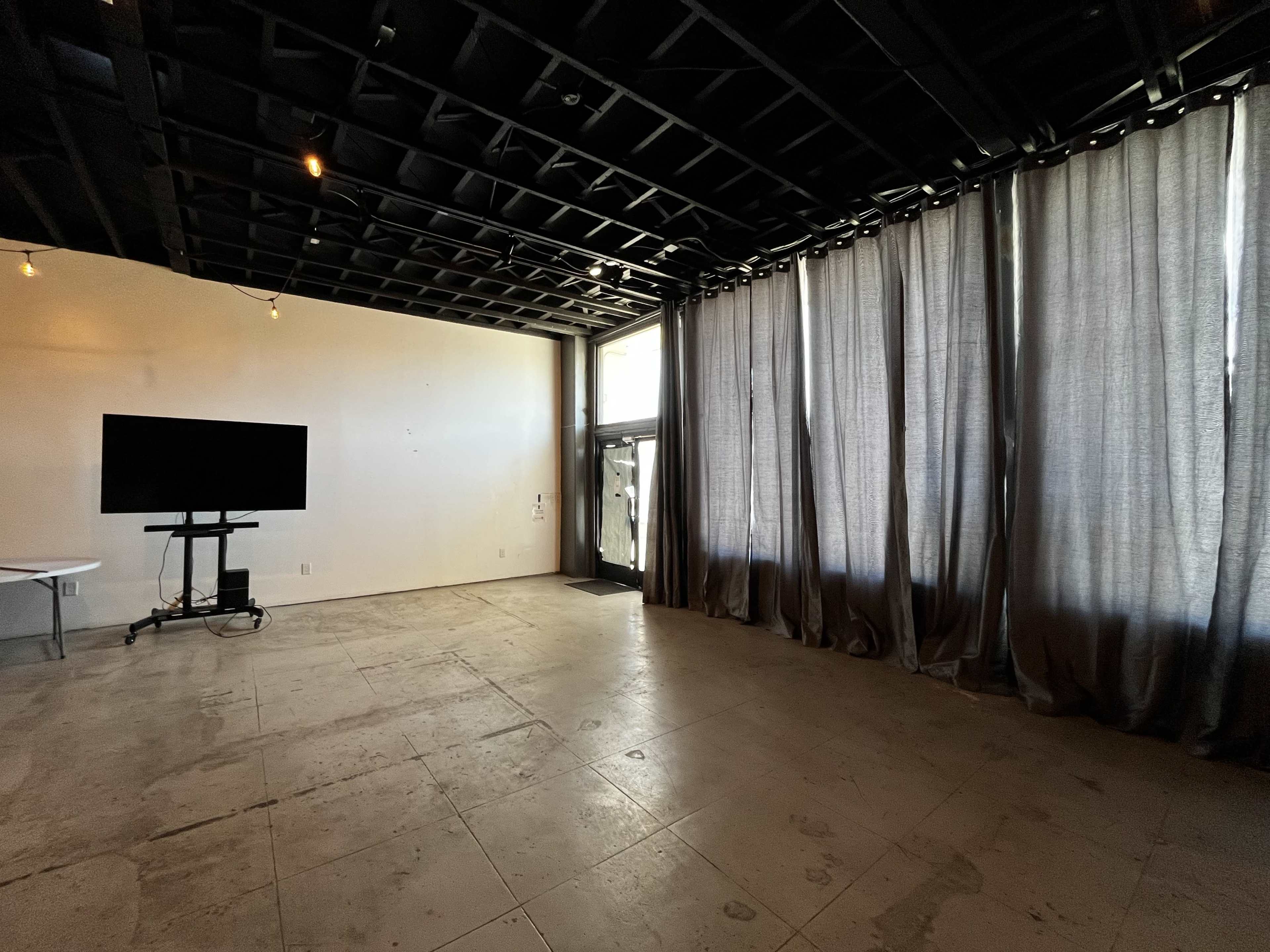 An empty room with a large window covered by dark curtains, a television mounted on a stand, and a concrete floor.