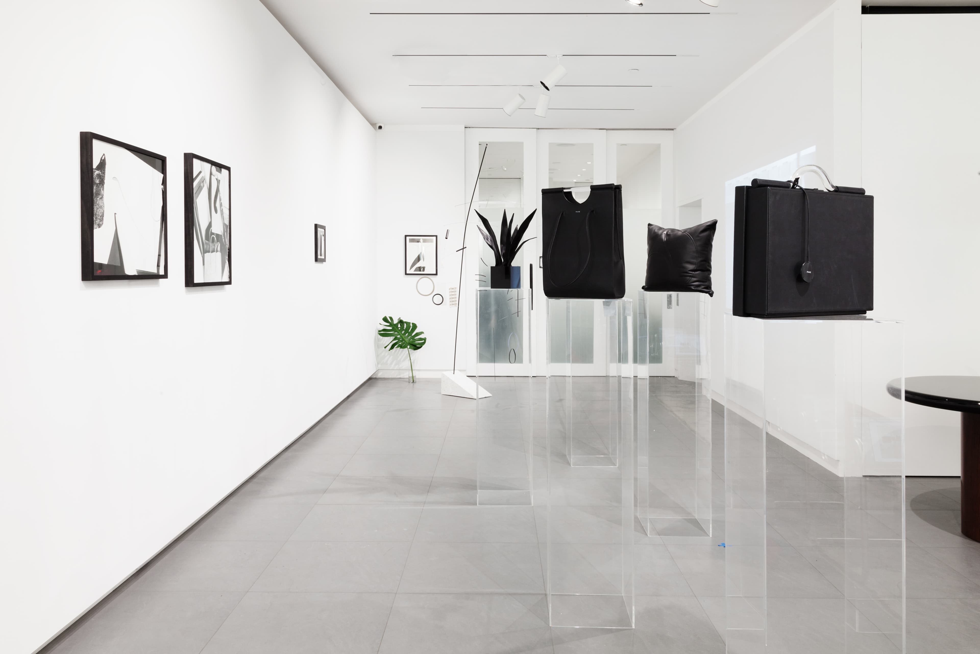The image shows a modern art gallery featuring two framed artworks on a white wall and three minimalist sculptures displayed on transparent pedestals.