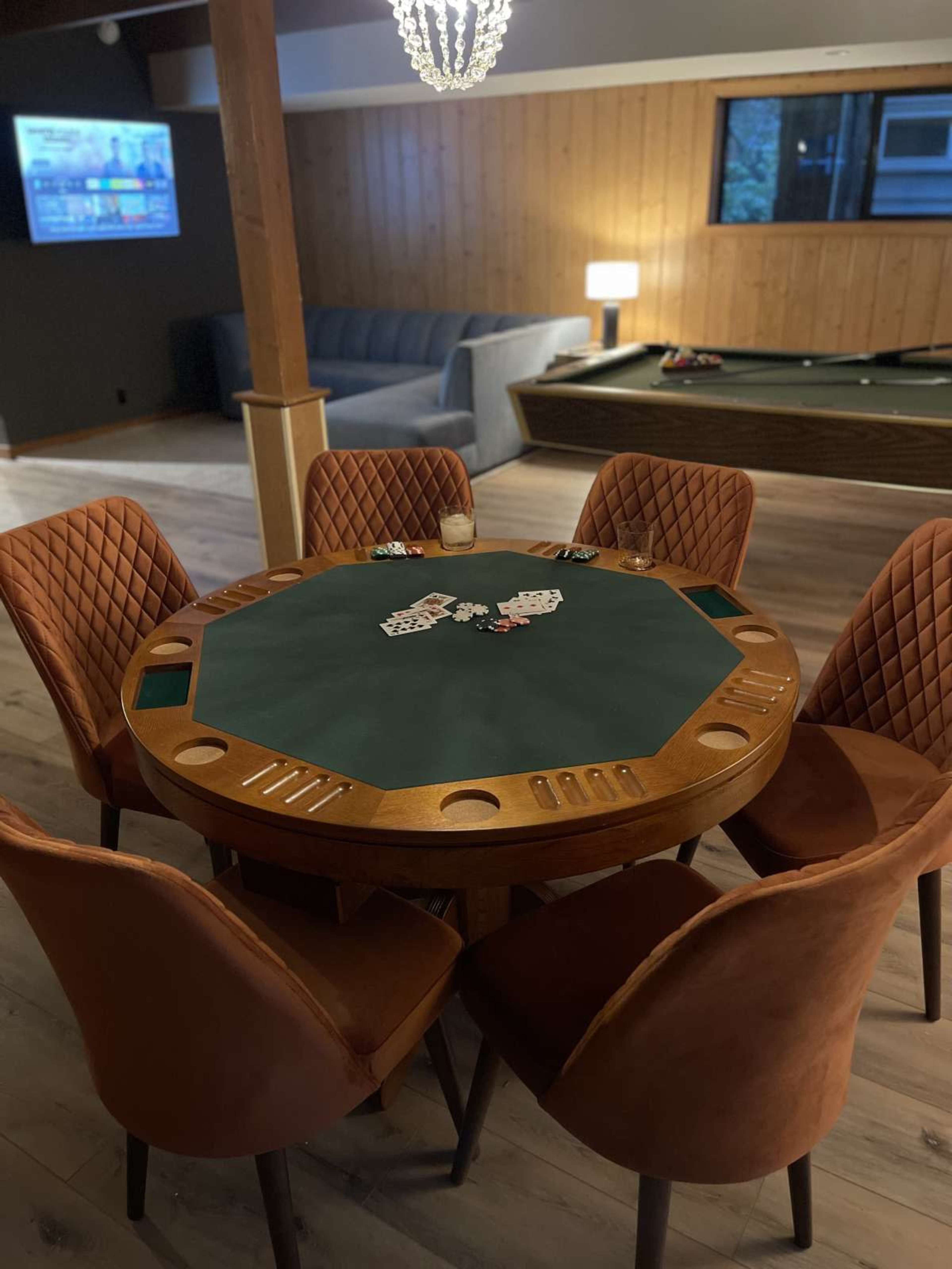 A round poker table with chips and cards is set up in a room featuring wooden paneling and a pool table in the background.