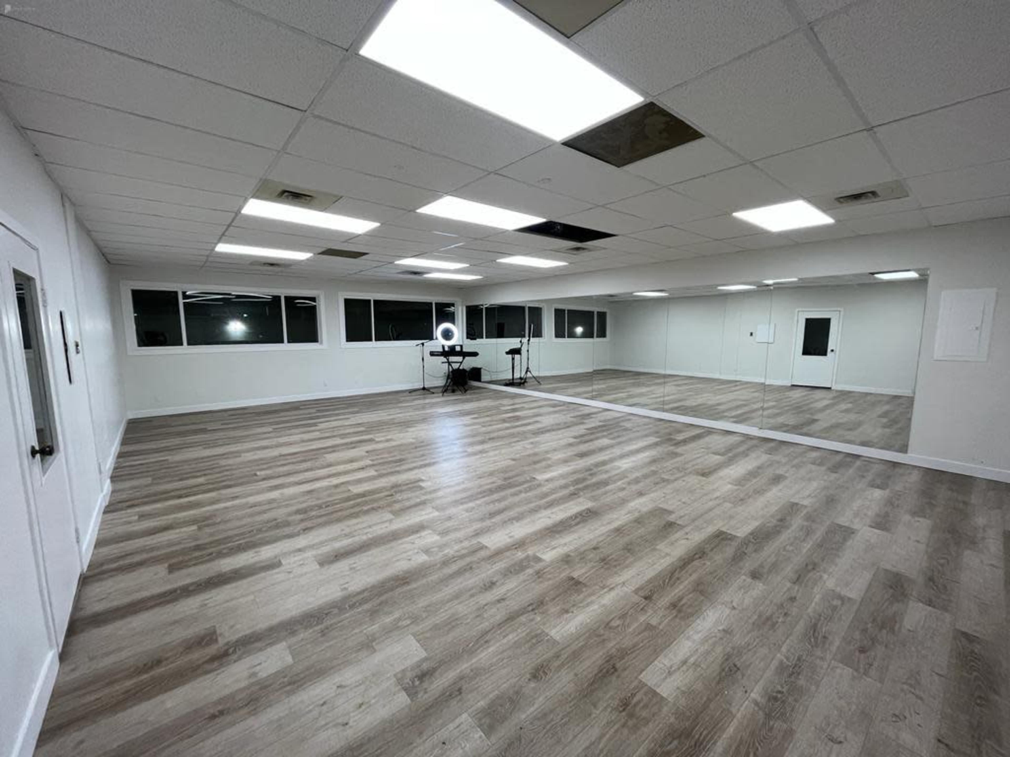 A spacious, empty studio room with a wooden floor, large windows, and a long mirror on one wall.