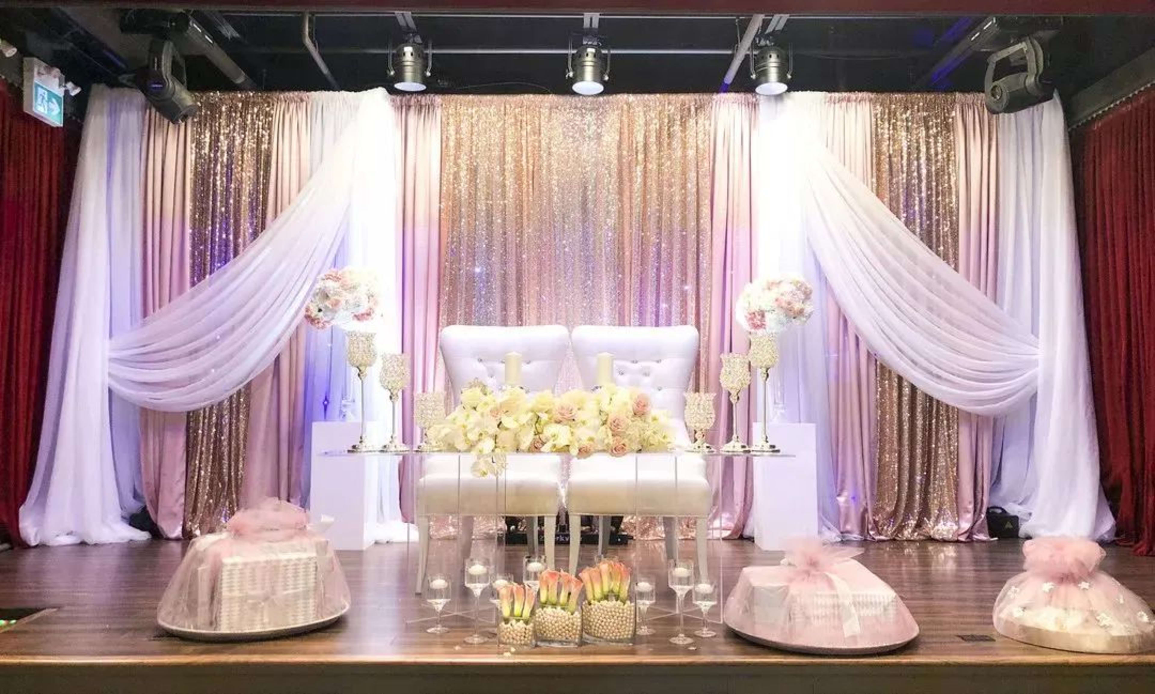 A staged event backdrop with a white sofa, floral arrangements, and draped fabric in shades of pink and gold.