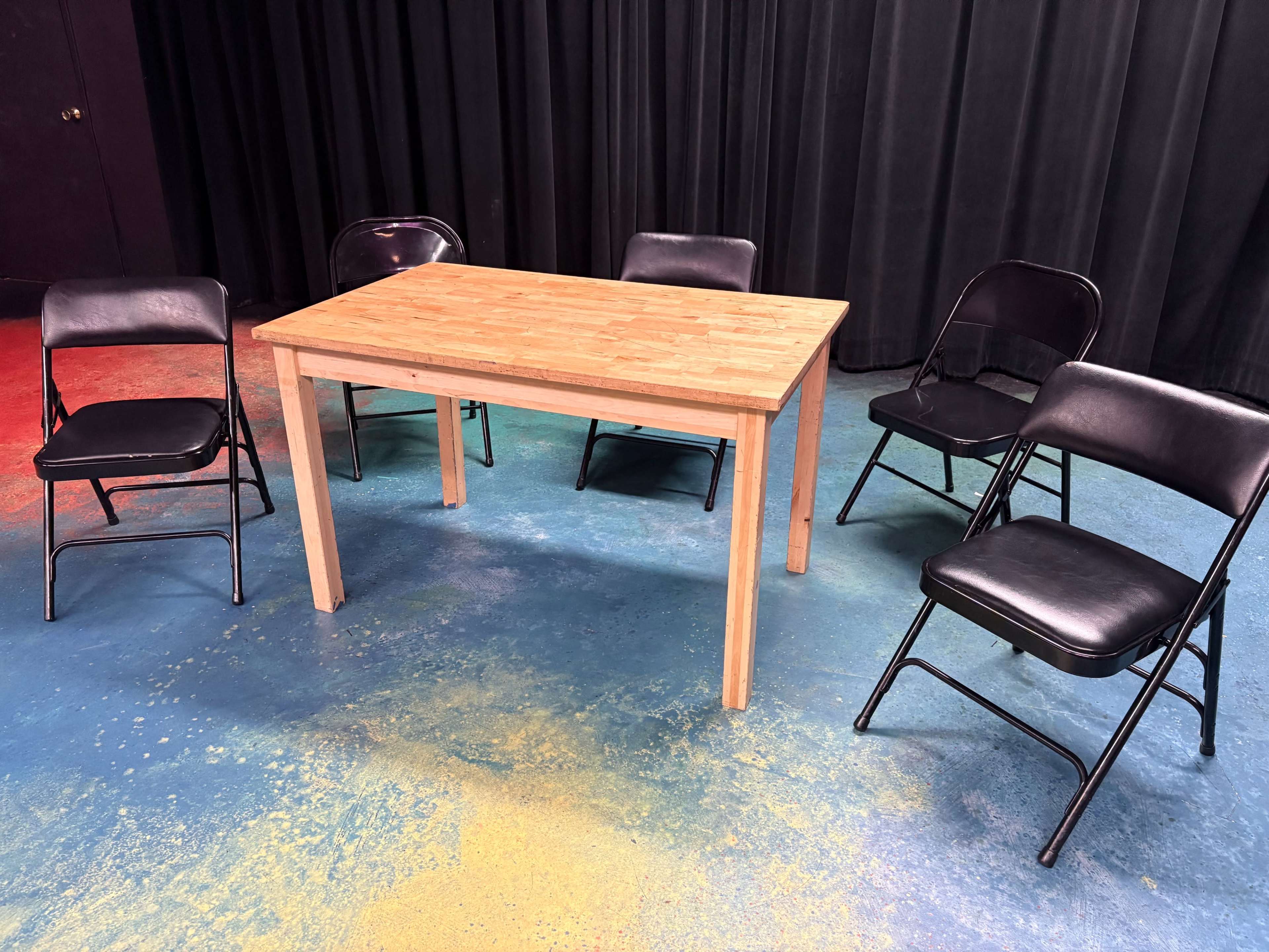 A wooden table surrounded by four black folding chairs on a colorful, splattered floor.