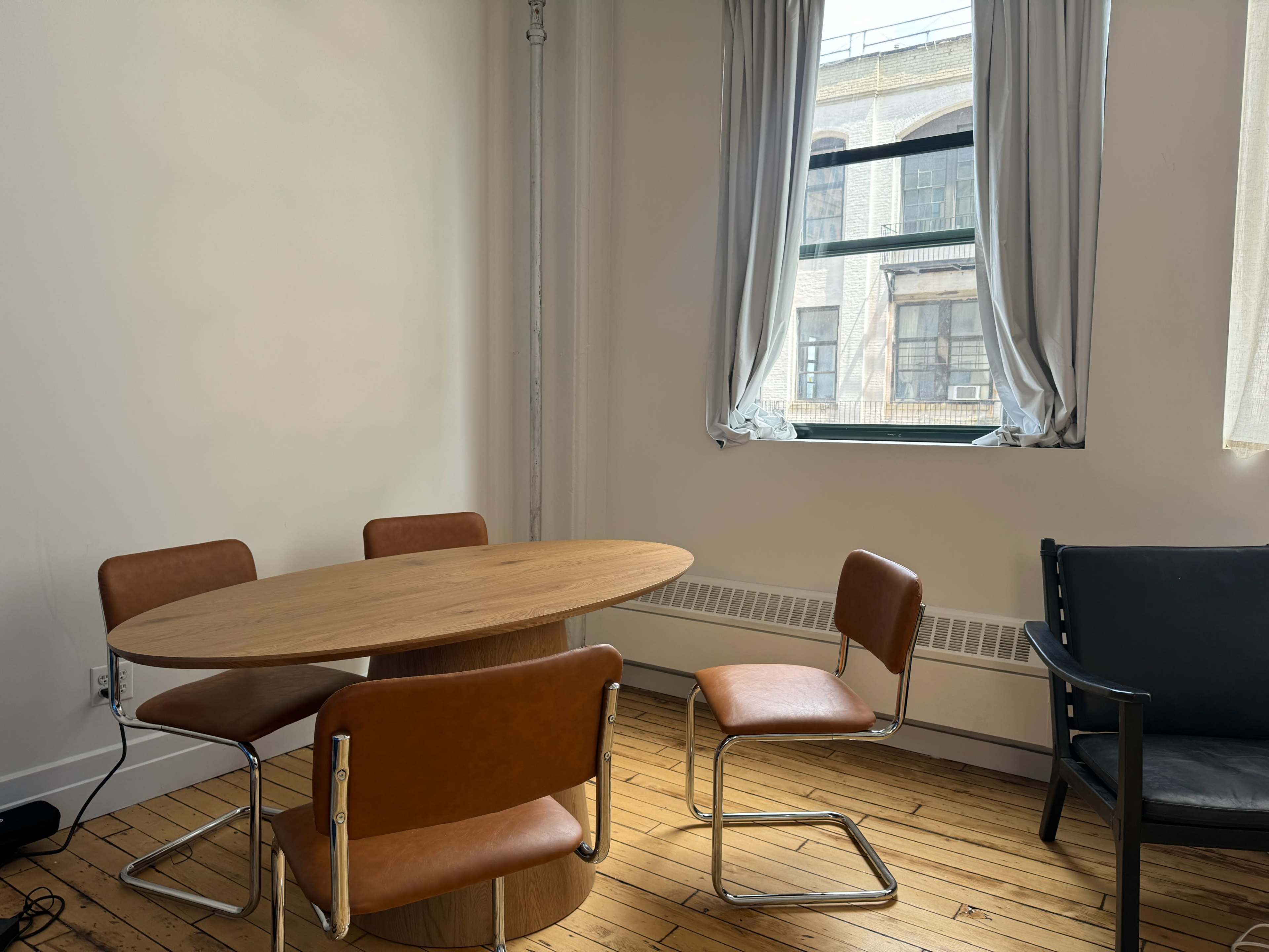 The image shows a small room featuring a round wooden table with four brown leather chairs and a large window with light-colored curtains.