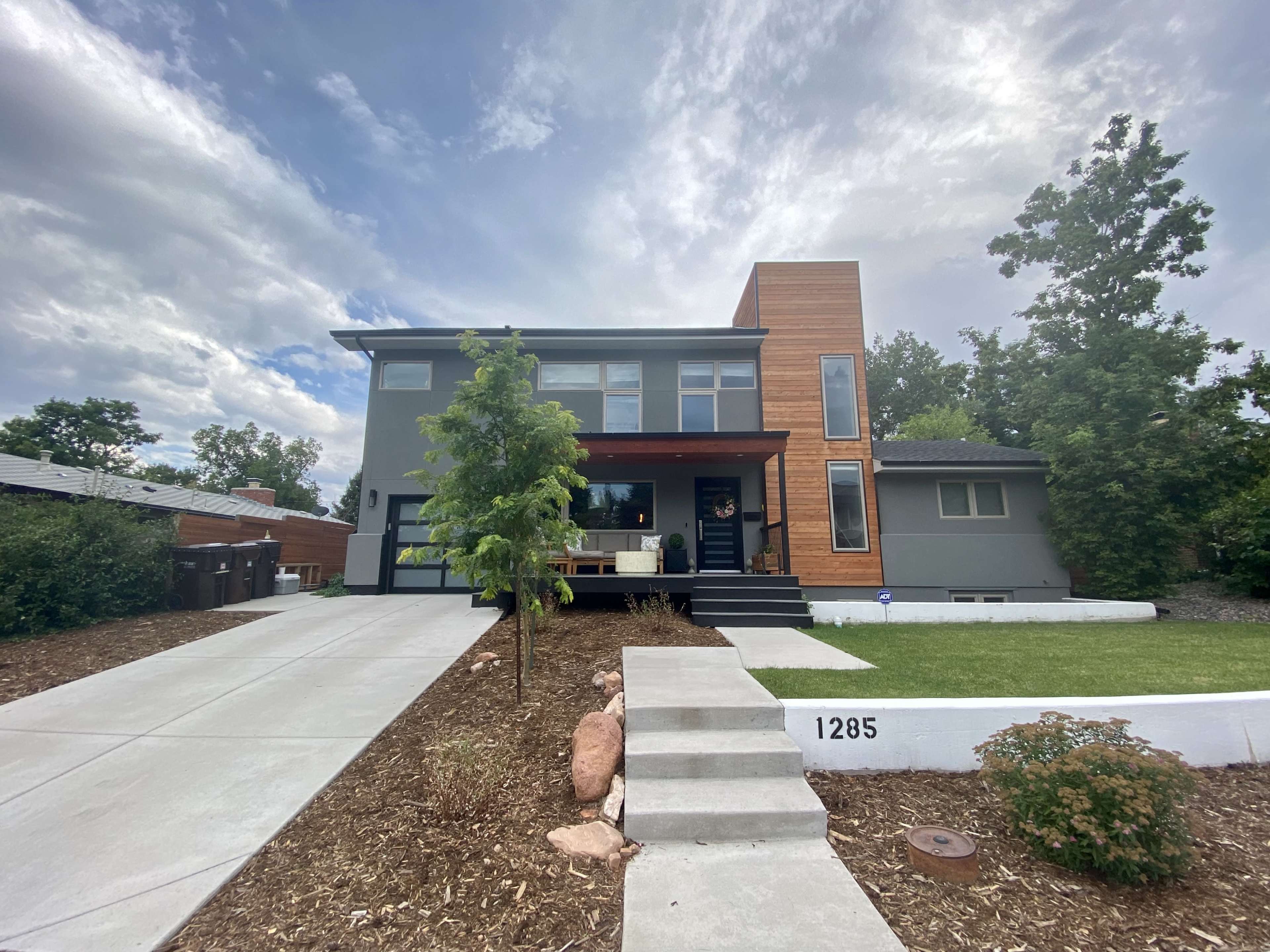 The image shows a modern two-story house with a mix of gray and wooden siding, featuring a landscaped front yard and a concrete driveway.