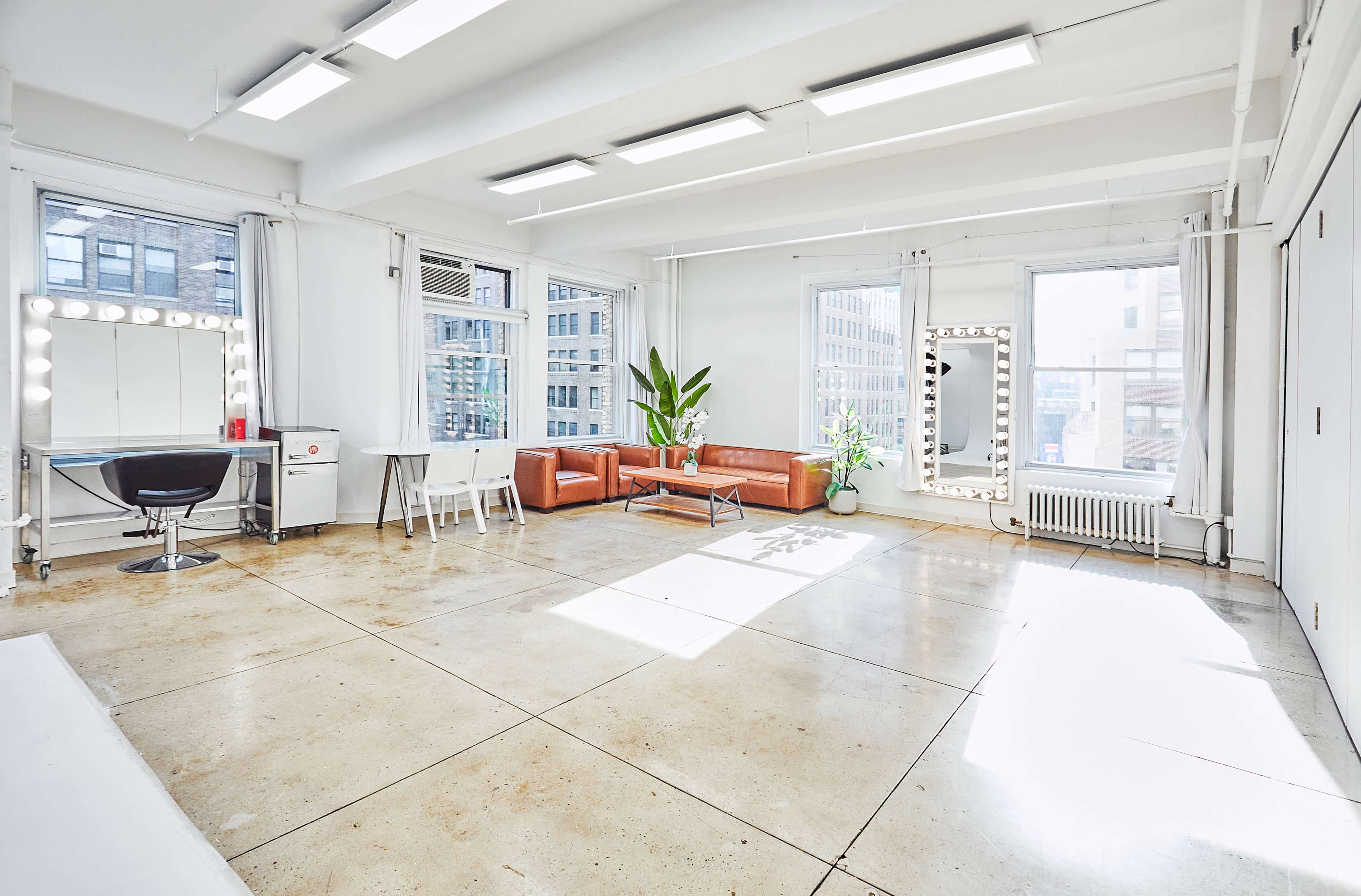 The image shows a spacious, well-lit room with large windows, featuring a seating area with brown couches, a white table, and a vanity mirror.