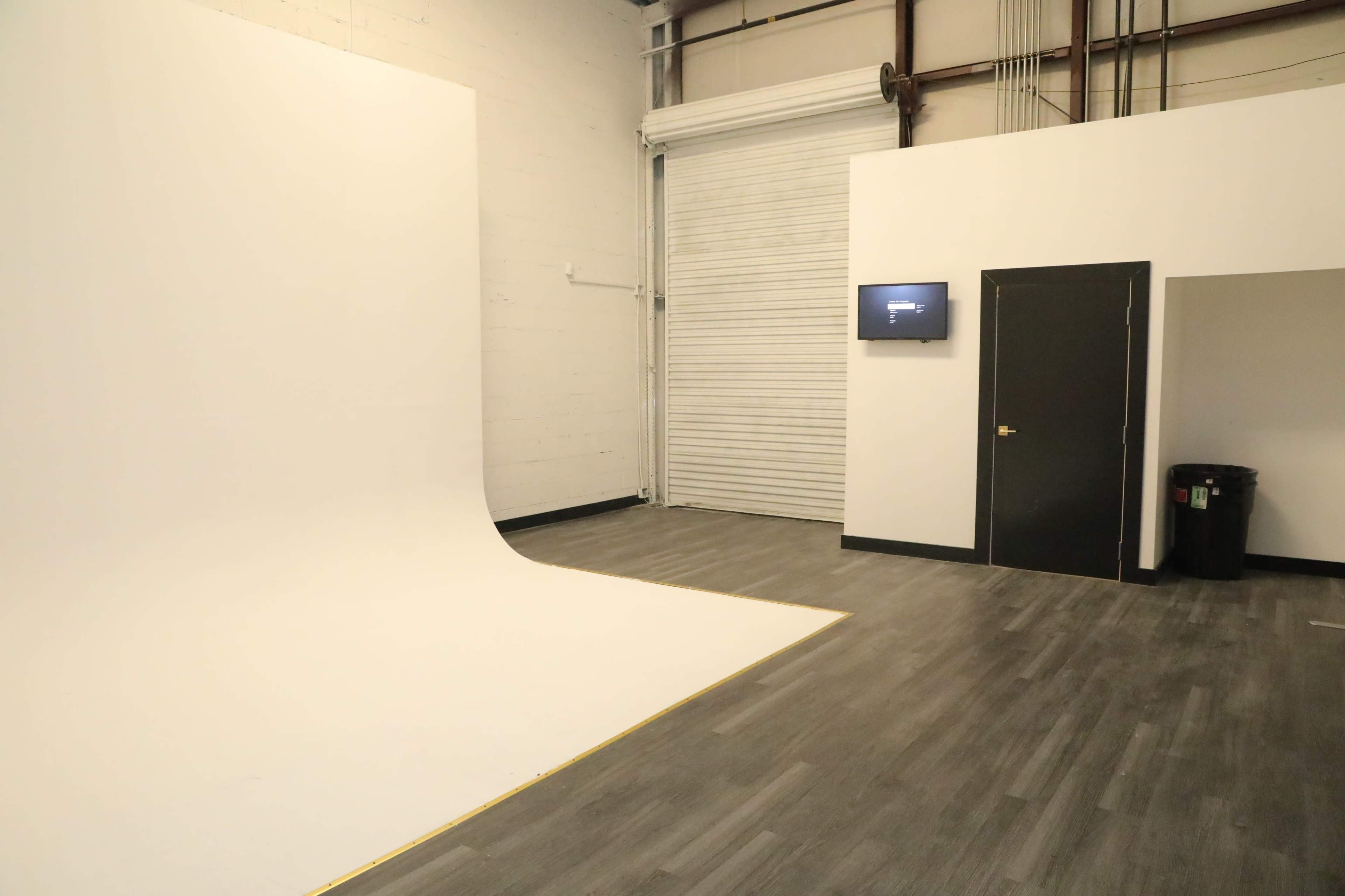 A photography studio with a smooth white backdrop, a closed door, a small monitor on the wall, and a trash can in the corner.