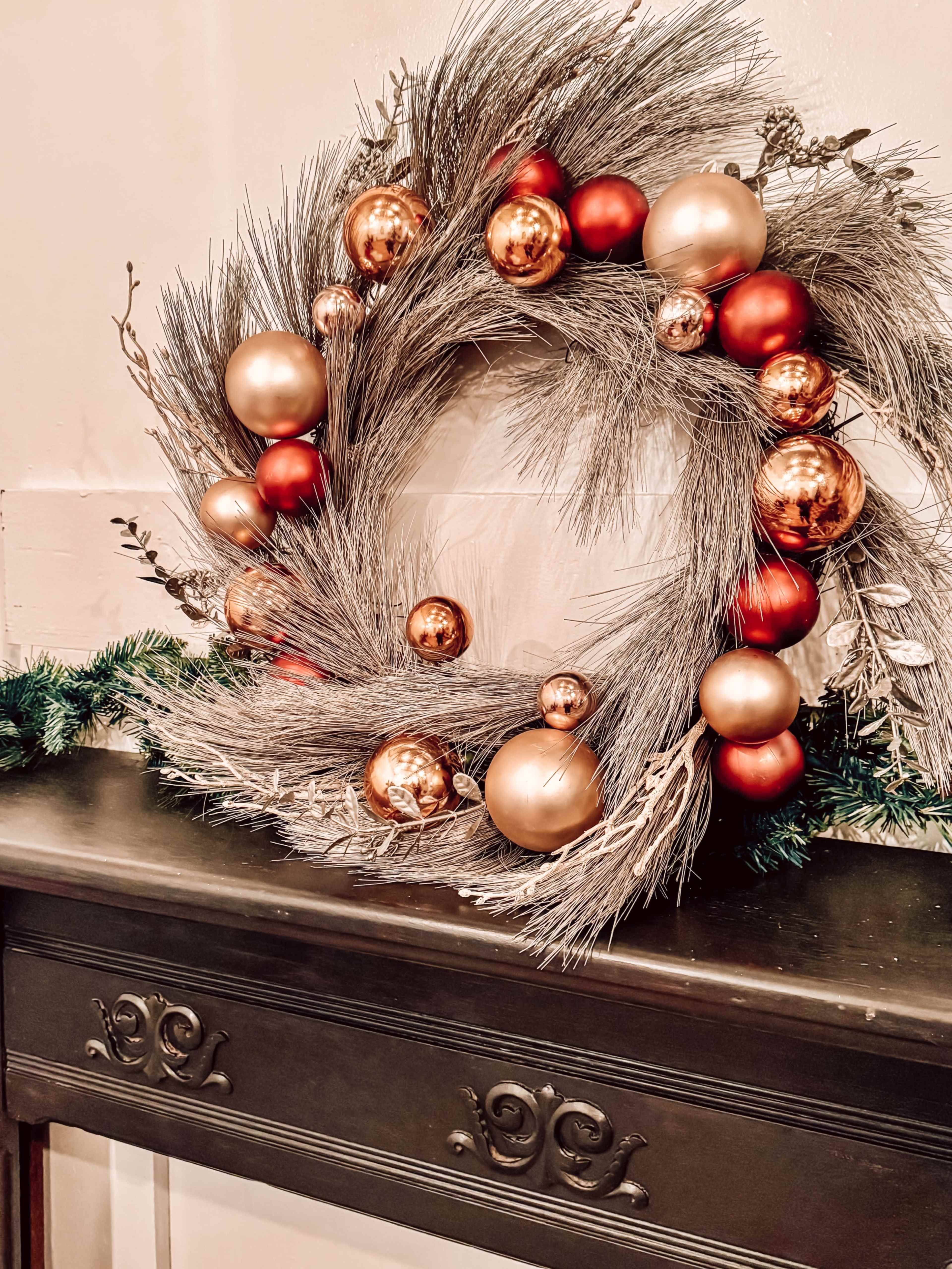 A decorative wreath made of silver foliage and adorned with red and gold baubles is displayed on a dark wooden mantel.