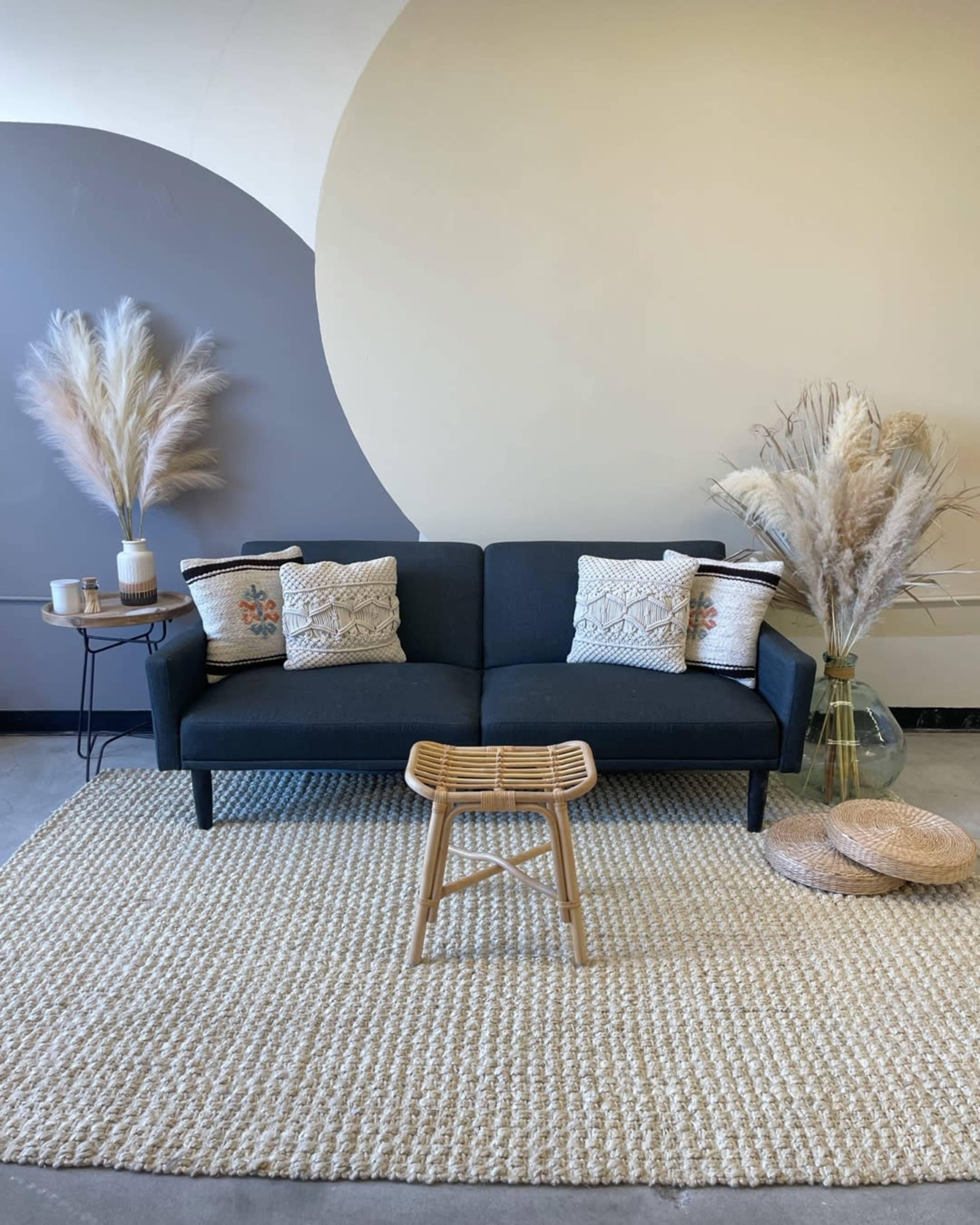 A dark sofa with decorative pillows is positioned on a textured rug, accompanied by a small stool and tall decorative grasses in vases.