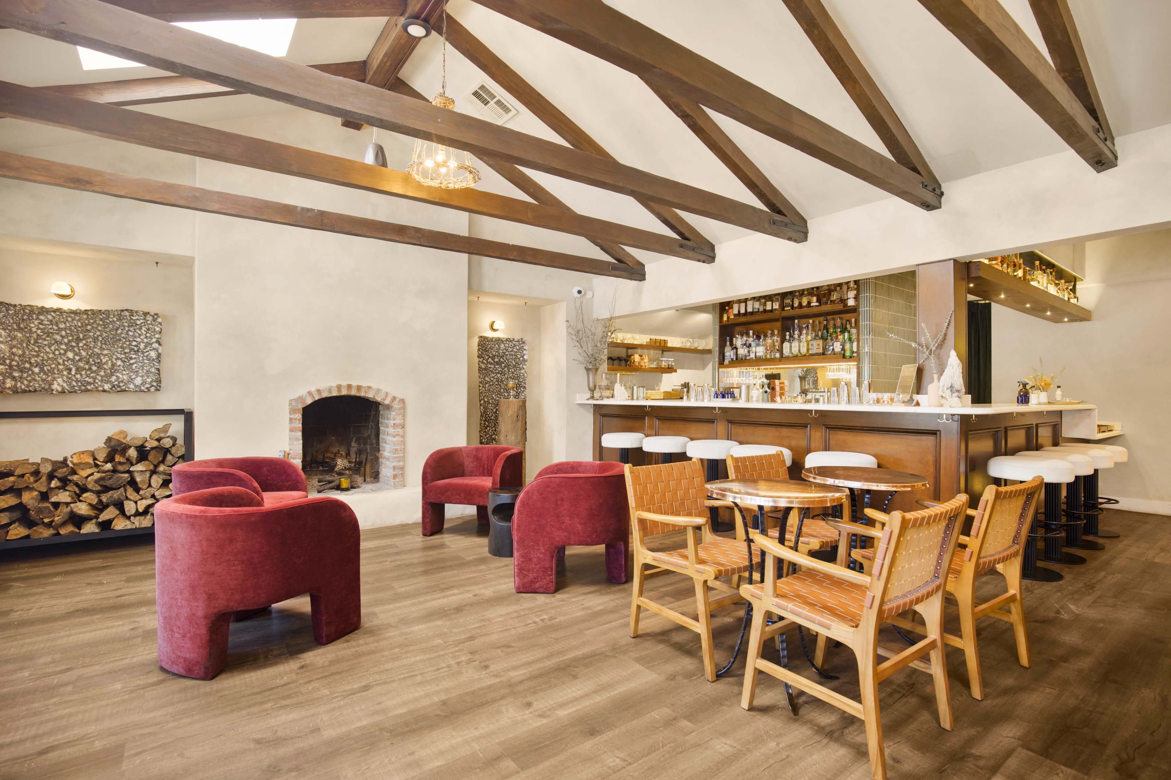 The image shows a modern bar area with wooden beams, a sleek counter, red upholstered chairs, and a cozy fireplace with stacked logs.