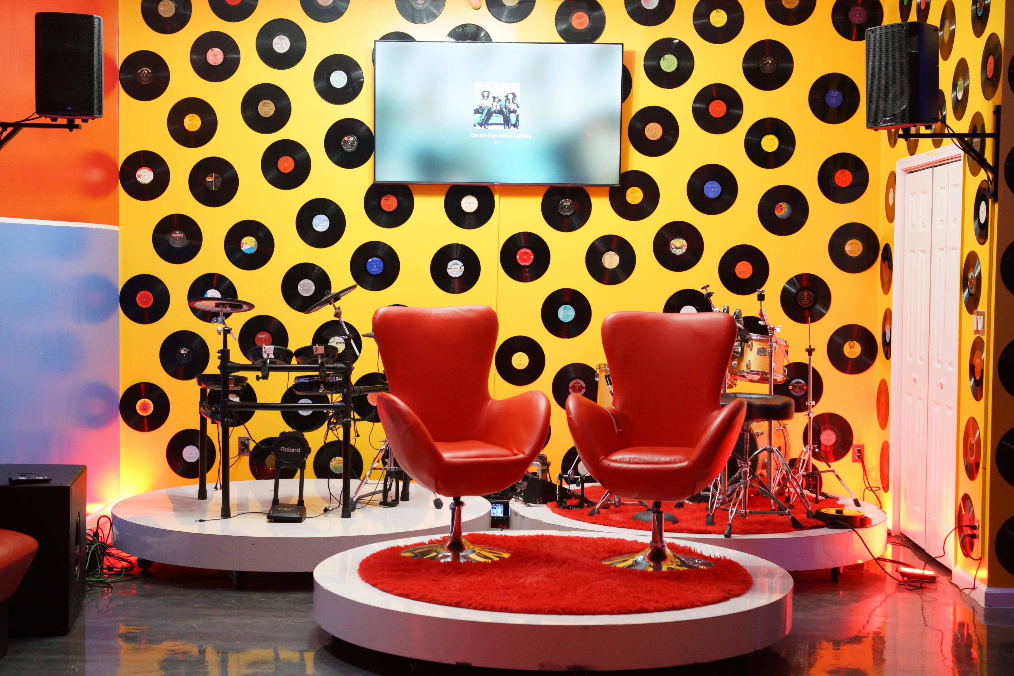 The image shows a vibrant room decorated with a yellow wall covered in black vinyl records, featuring two red chairs on a circular platform, a drum set, and a television mounted above.
