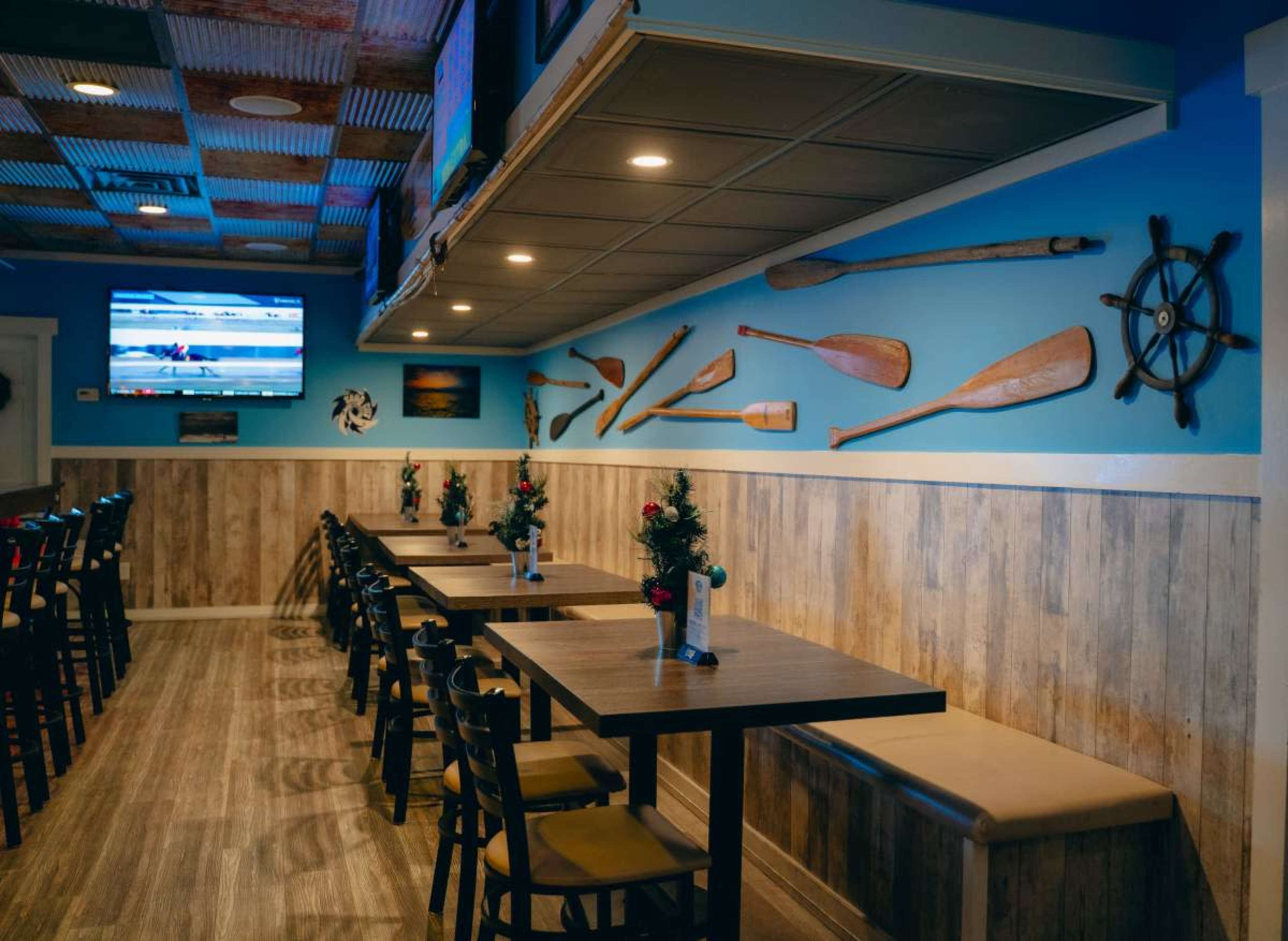 The image shows a restaurant interior with wooden tables, black chairs, and decorative paddles on the blue wall, alongside a television mounted above.