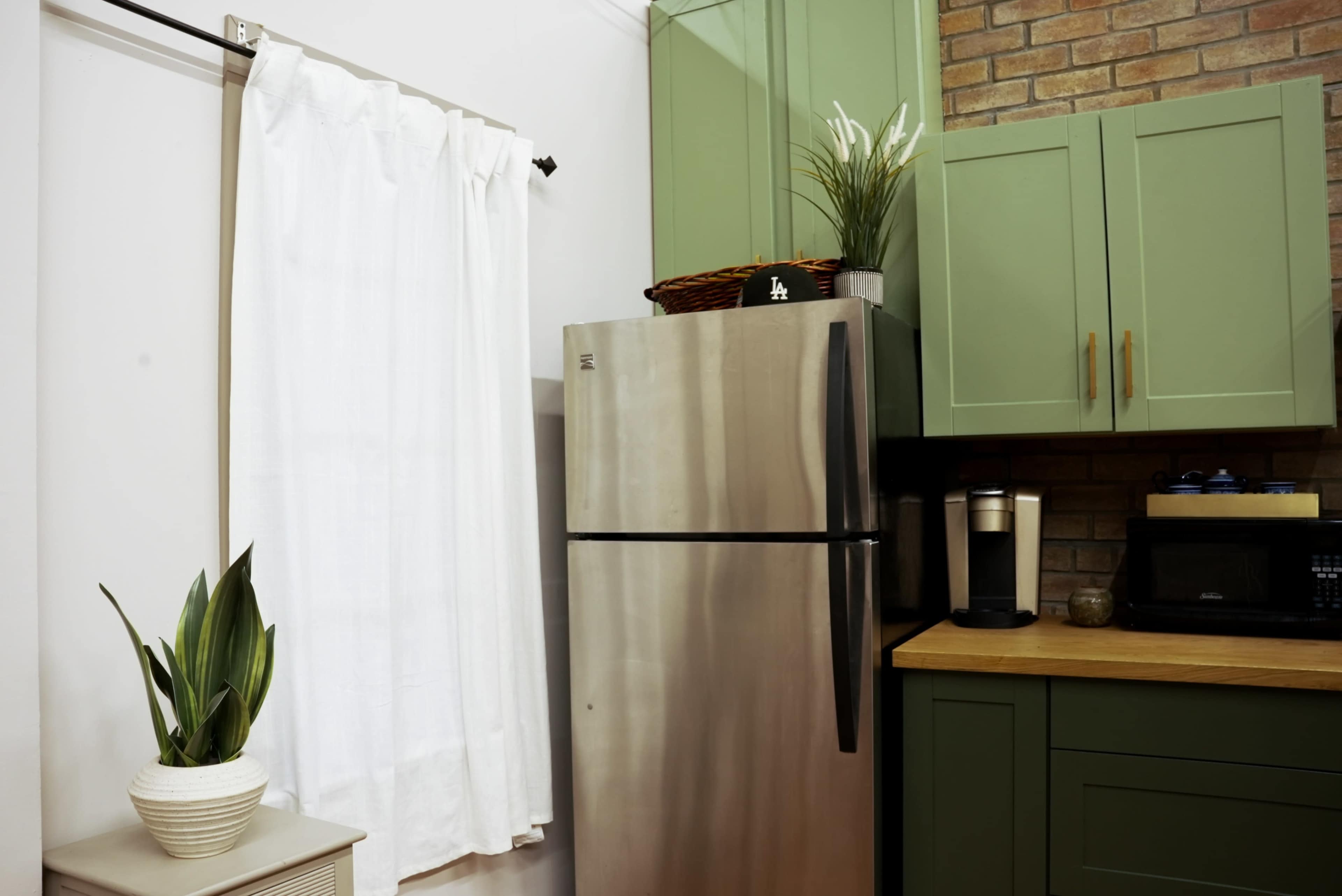 The image shows a kitchen corner with a stainless steel refrigerator, green cabinetry, a countertop coffee maker, and a white curtain covering a window.