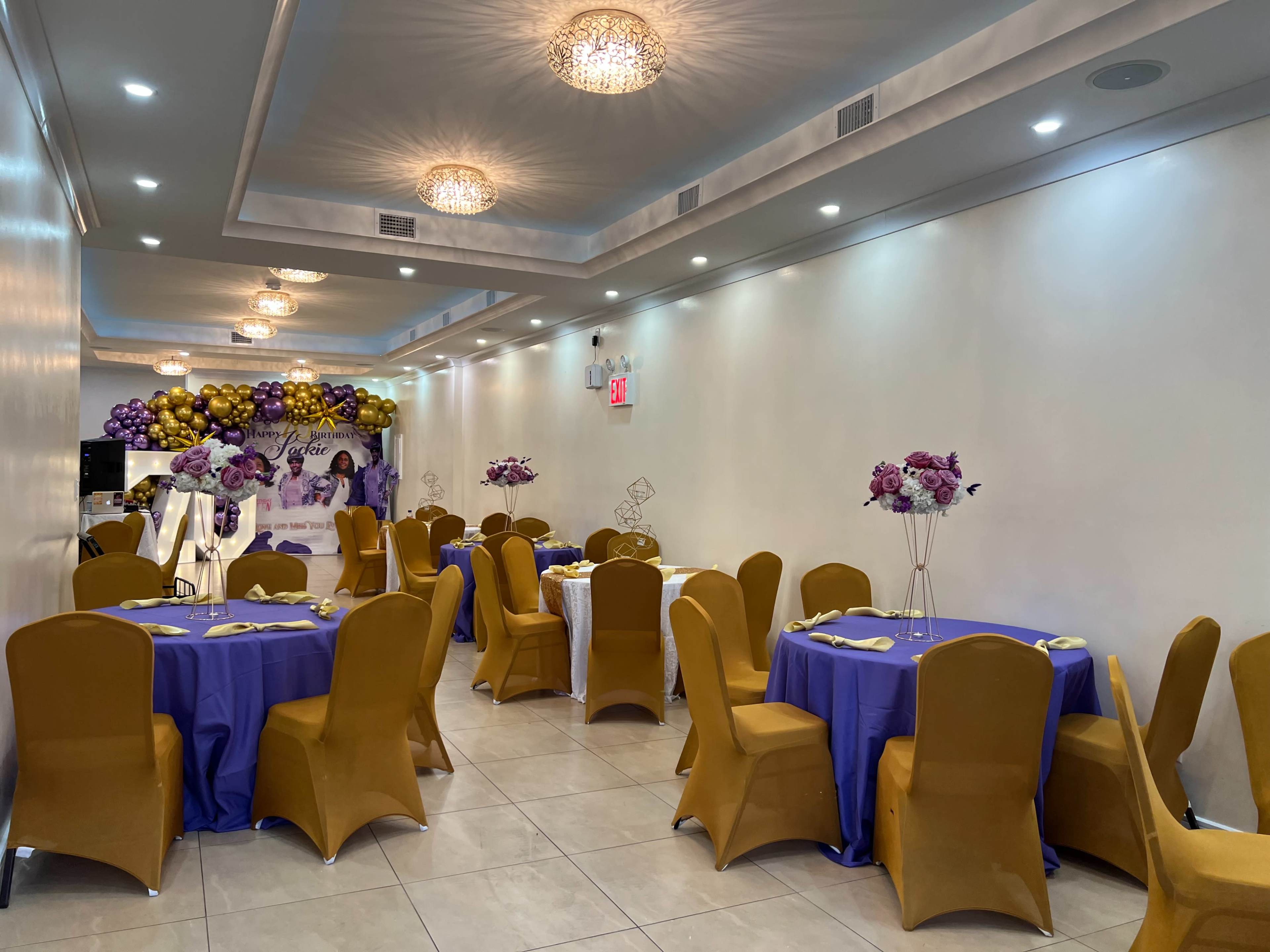 A well-lit event space features tables dressed in purple and gold linens, adorned with floral centerpieces.