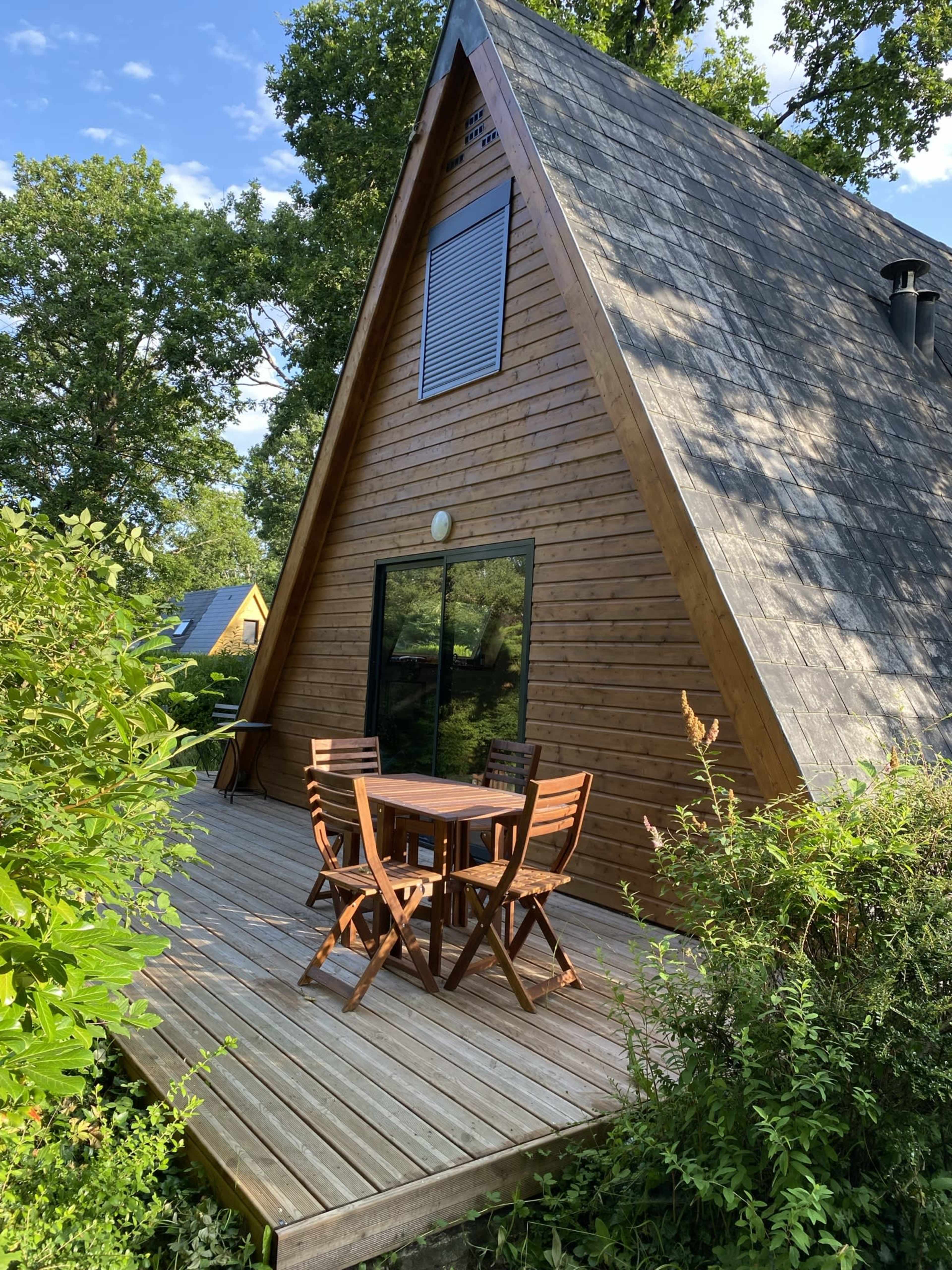 A wooden A-frame cabin with a large glass front and a deck featuring a table and chairs is surrounded by lush greenery.
