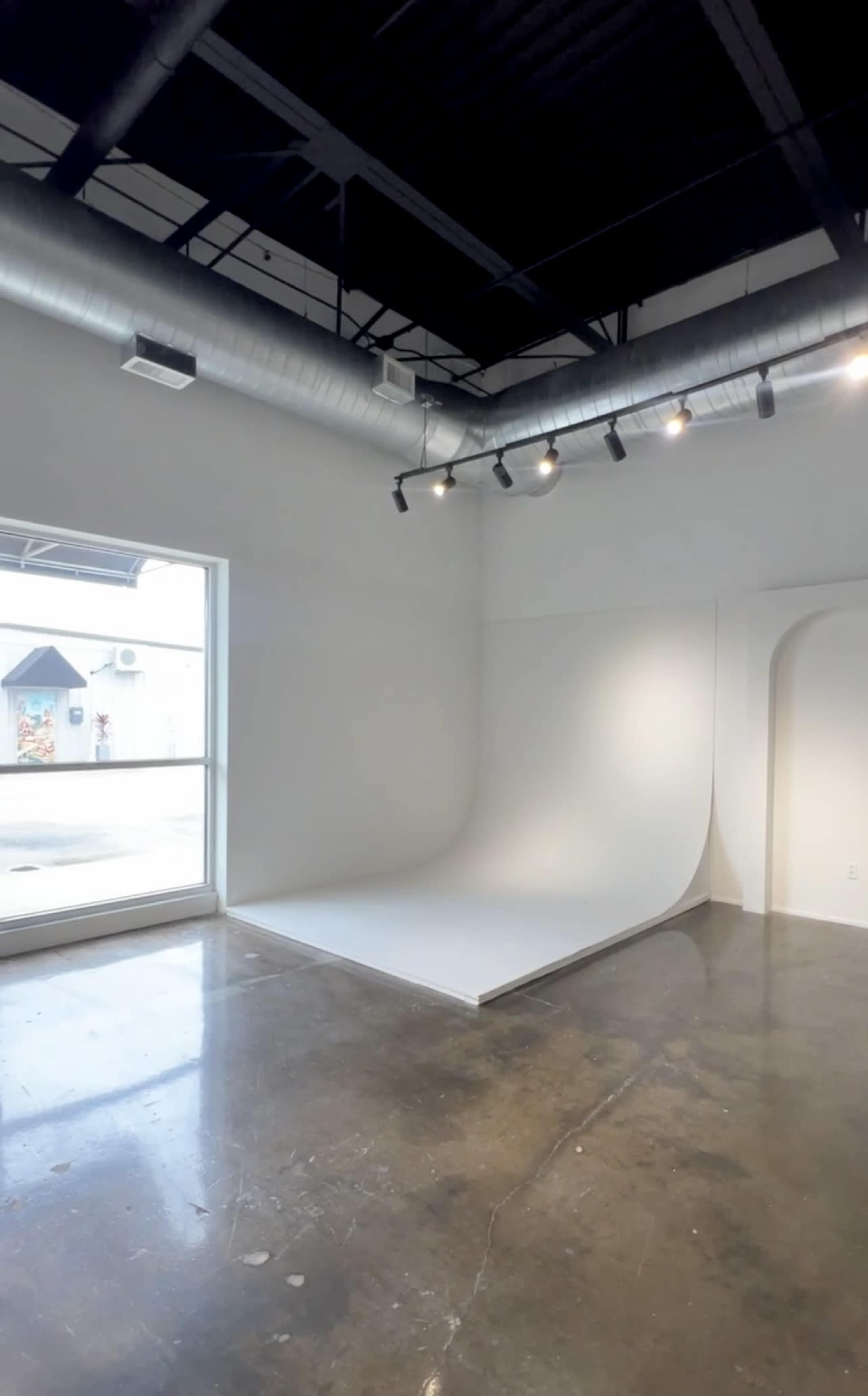 The image shows a spacious, empty studio with a large window, a seamless white backdrop, and polished concrete flooring.