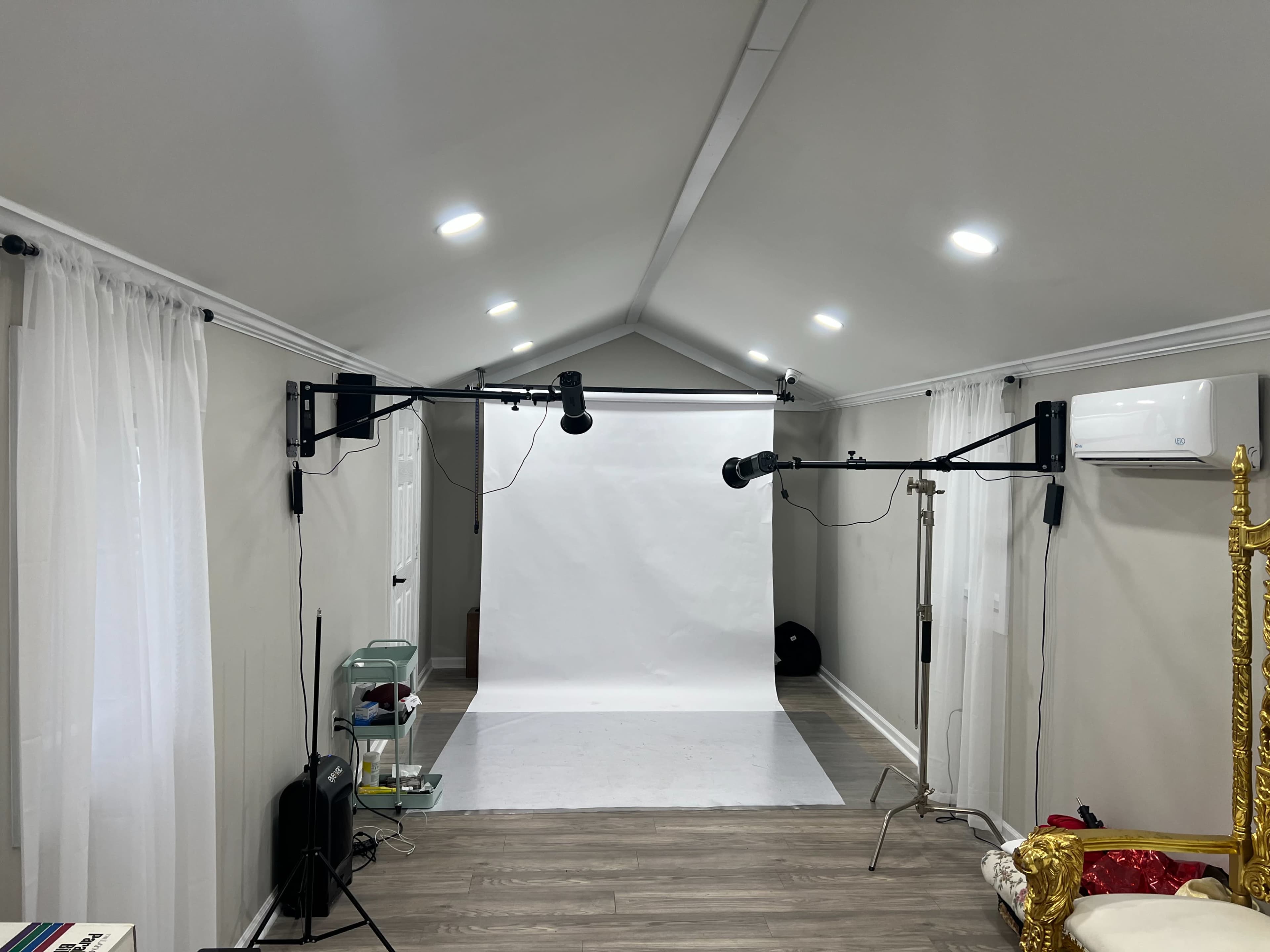 The image shows a bright photography studio with white walls, a large white backdrop in the center, and various lighting equipment mounted on black arms.