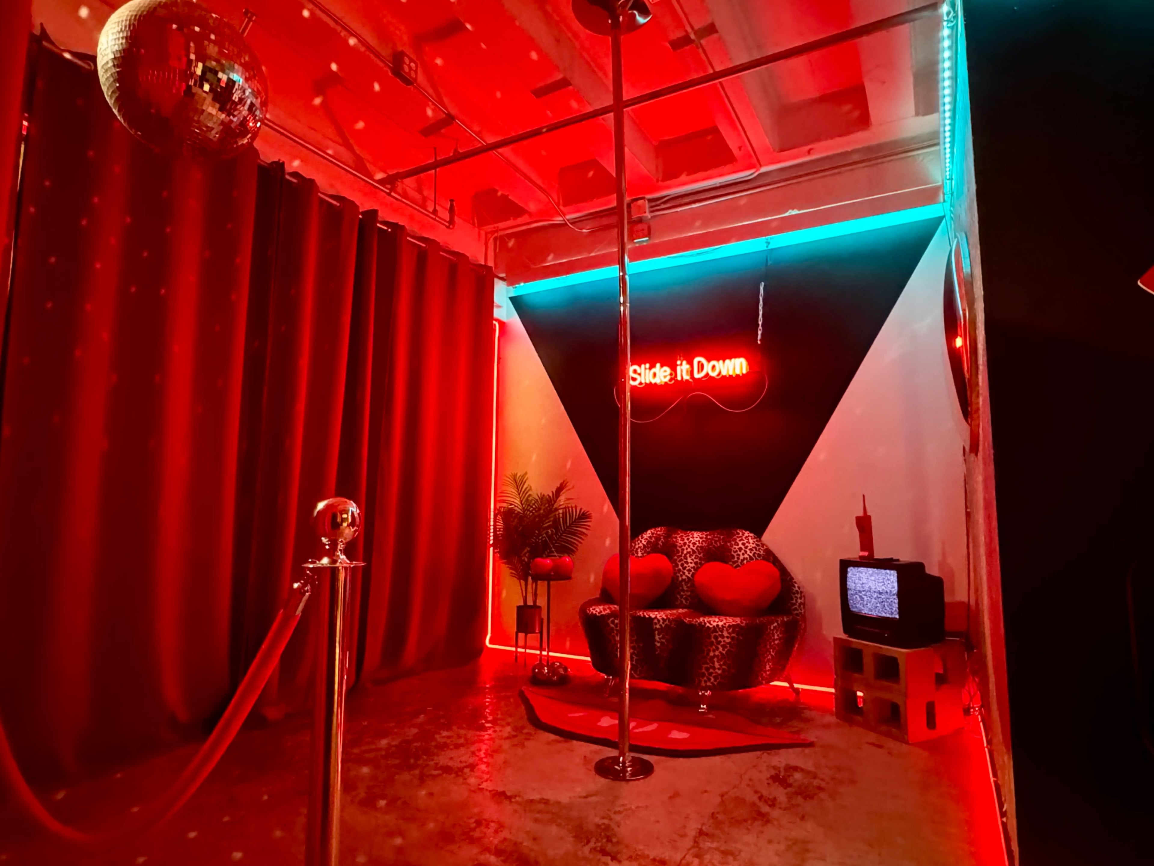 The image shows a dimly lit room with red lighting, featuring a velvet couch, a pole in the center, a retro TV, and a sign that says "Slide it Down."