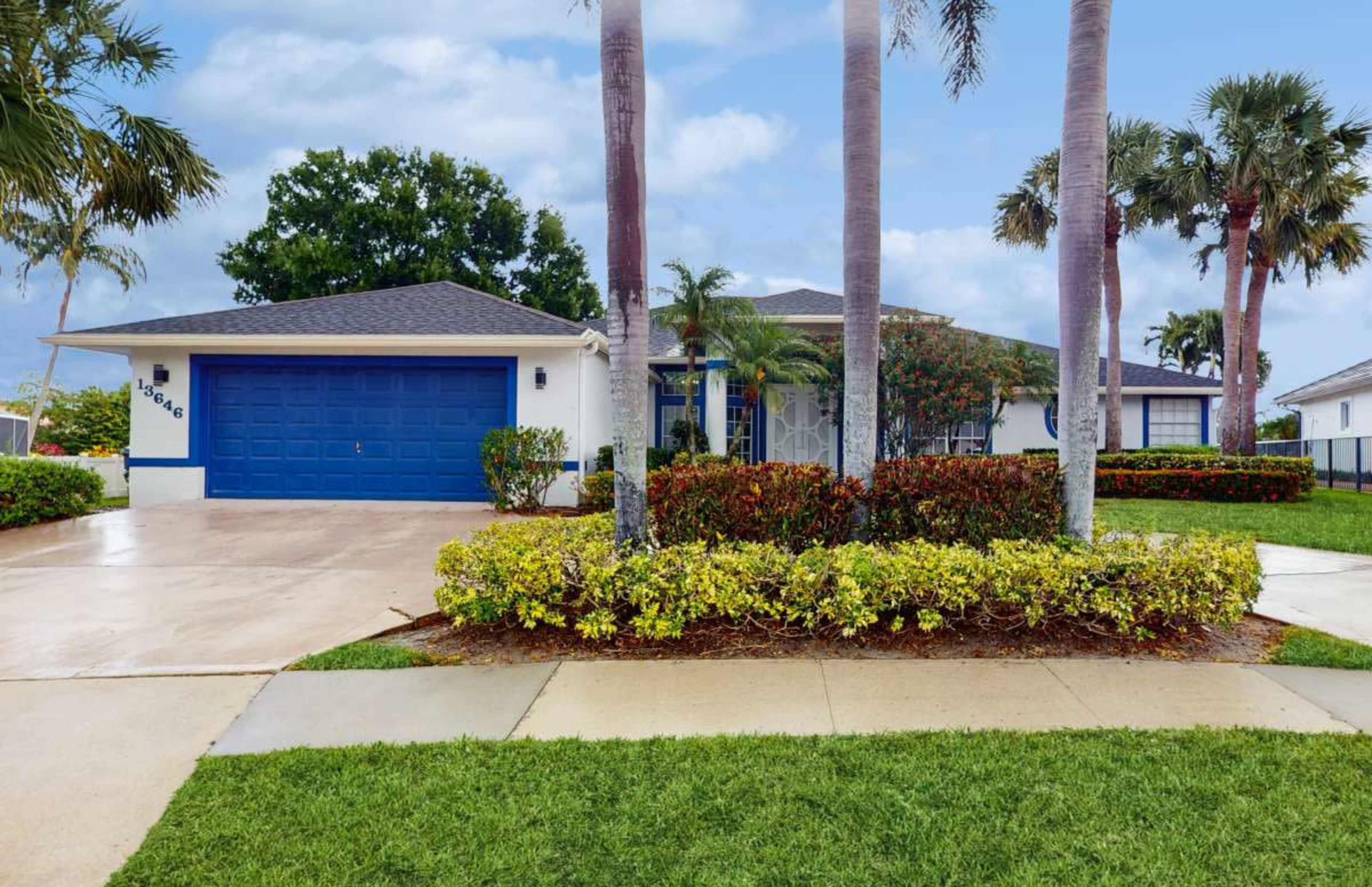 The image shows a single-story house with a blue garage door, a well-maintained lawn, and palm trees surrounding the property.