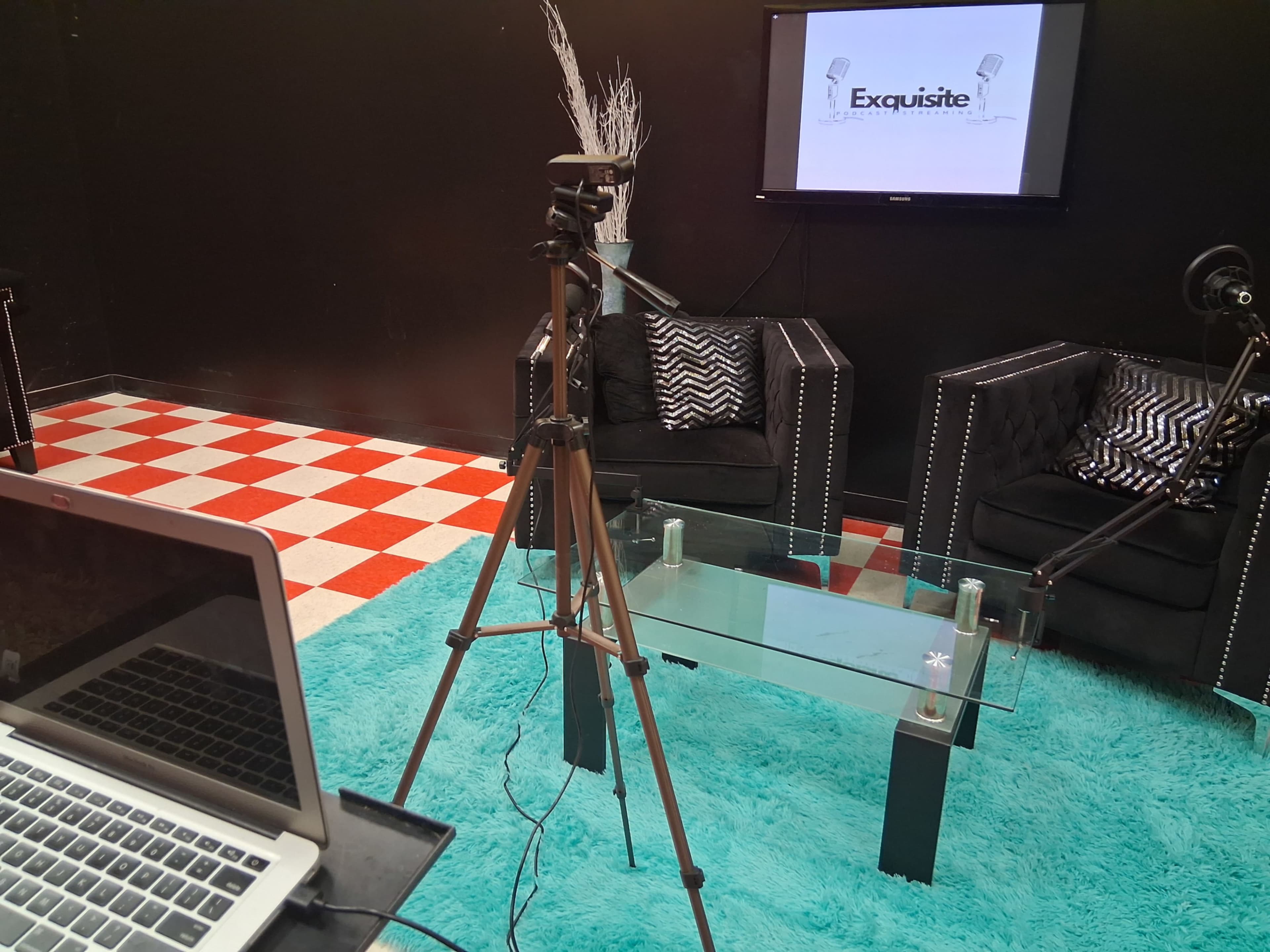 The image shows a recording studio setup with a tripod, a laptop on a table, and two black chairs positioned around a glass table on a light blue carpet.