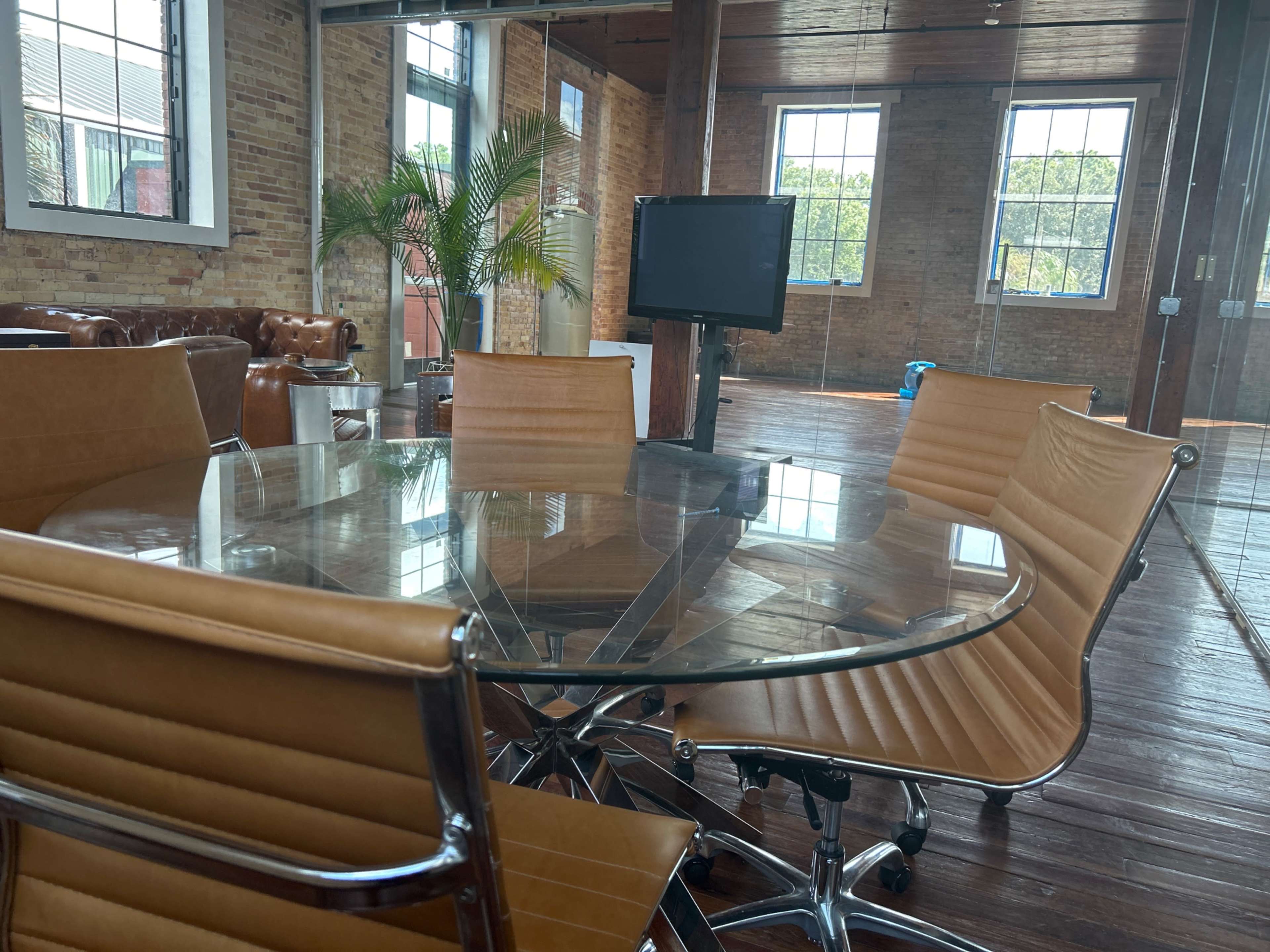 A glass conference table surrounded by four tan chairs is positioned in a well-lit room with large windows and exposed brick walls.