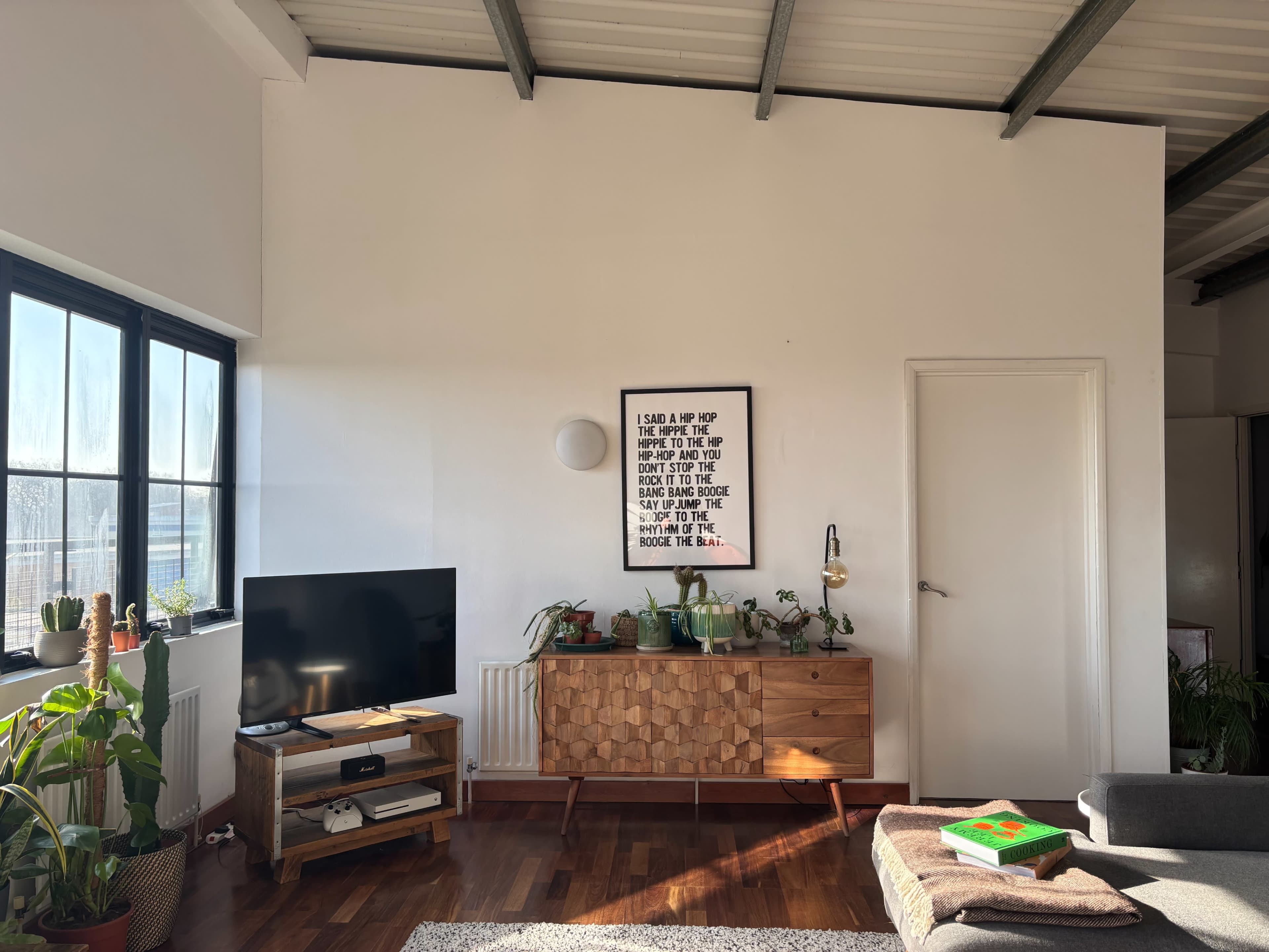 The image shows a modern living room featuring a television on a wooden stand, a stylish plant display, a wall-mounted light, and a framed quote centered above a wooden sideboard.