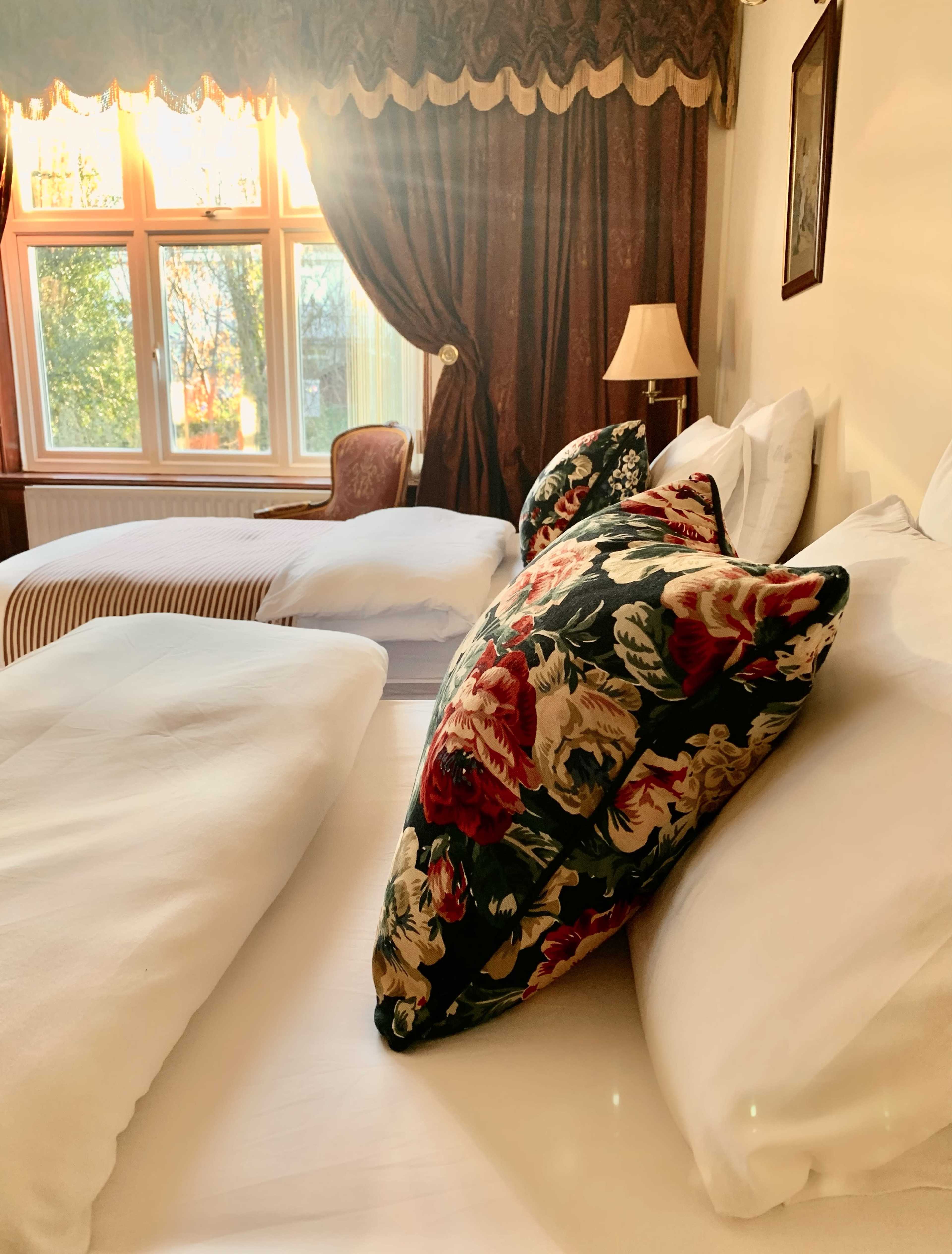 A well-lit hotel room with two beds, one adorned with a floral pillow, and a window allowing sunlight to stream in.