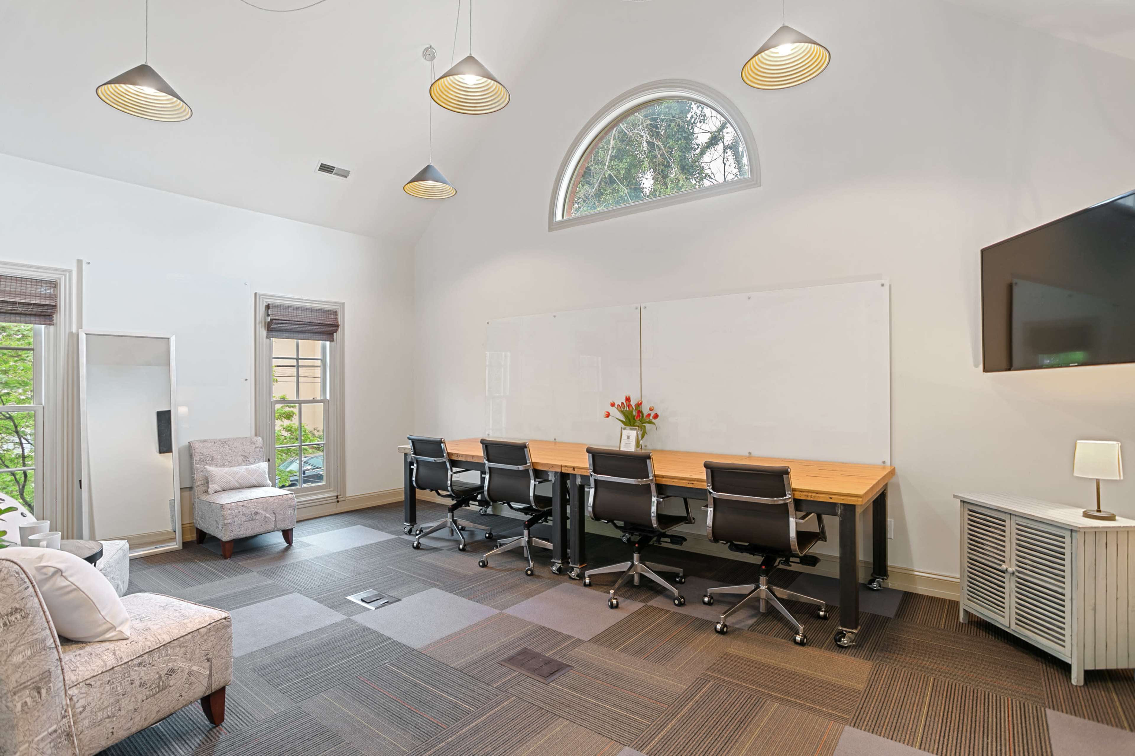 A modern meeting room features a large wooden table with four black chairs, a comfortable armchair, and a wall-mounted TV, all set under arched windows and pendant lighting.