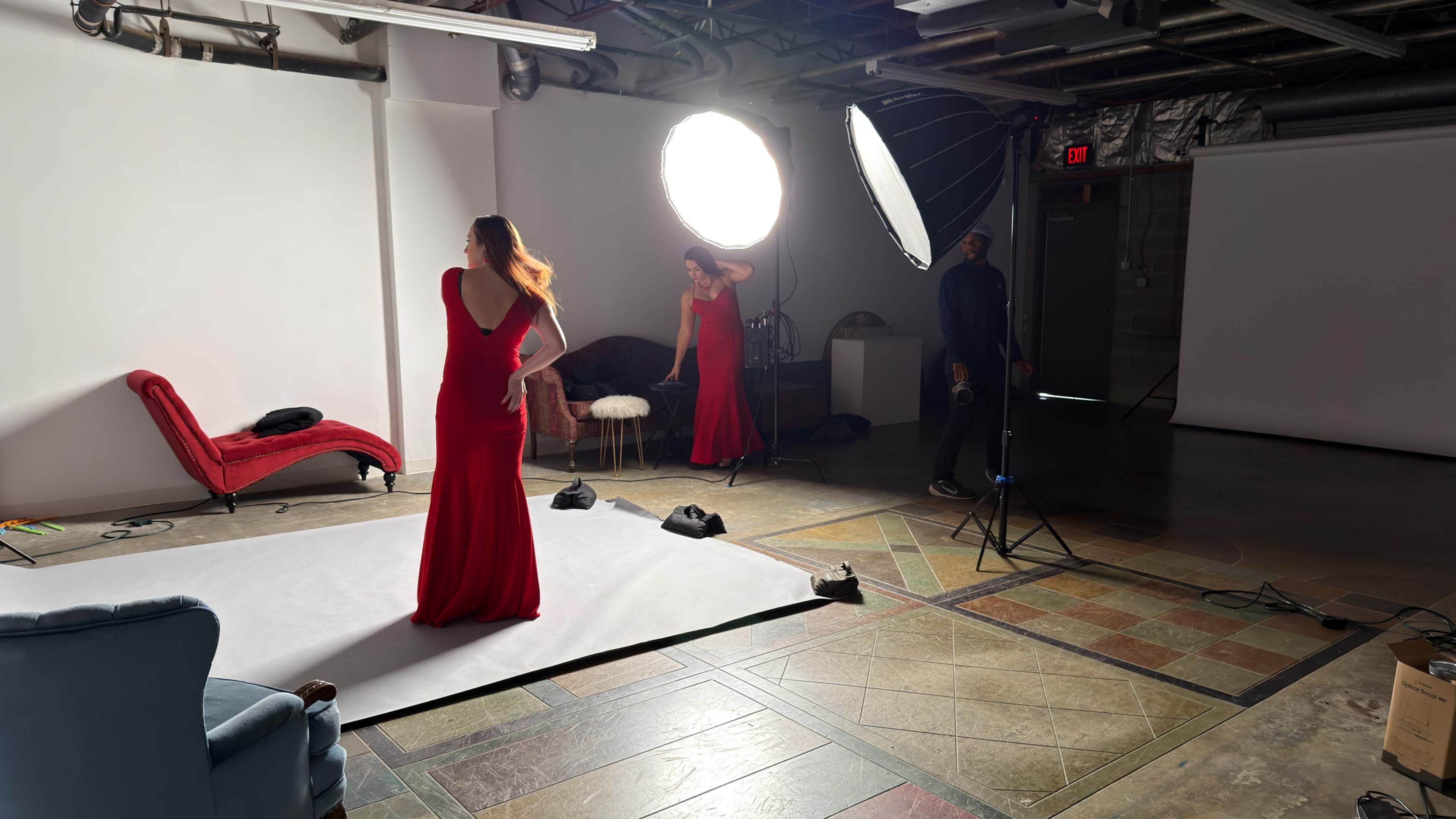 A model in a red dress poses in a photography studio, with a large light source illuminating the scene while a photographer captures the shot.