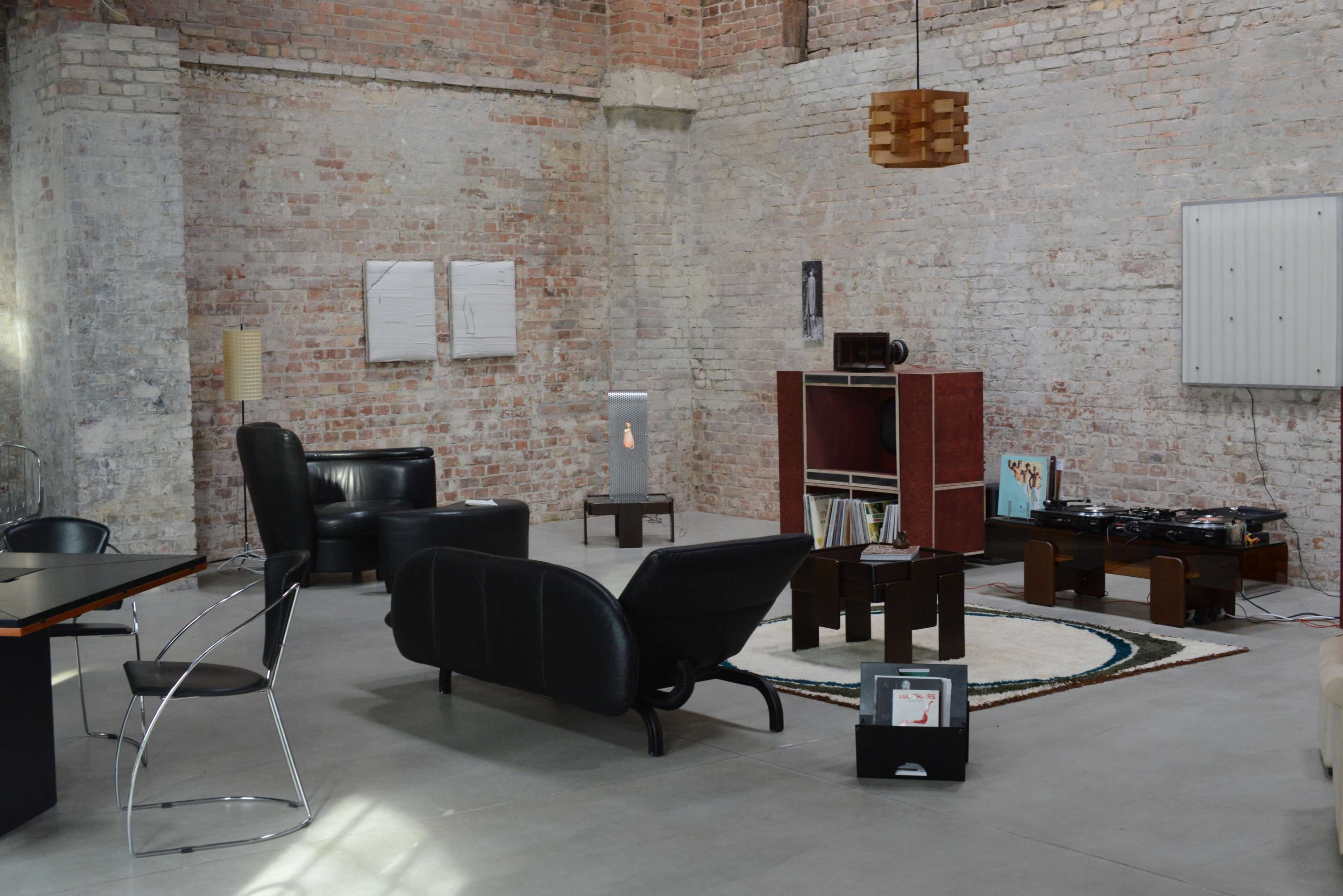 The image shows a minimalist living space featuring black leather furniture, a wooden bookshelf, and exposed brick walls.