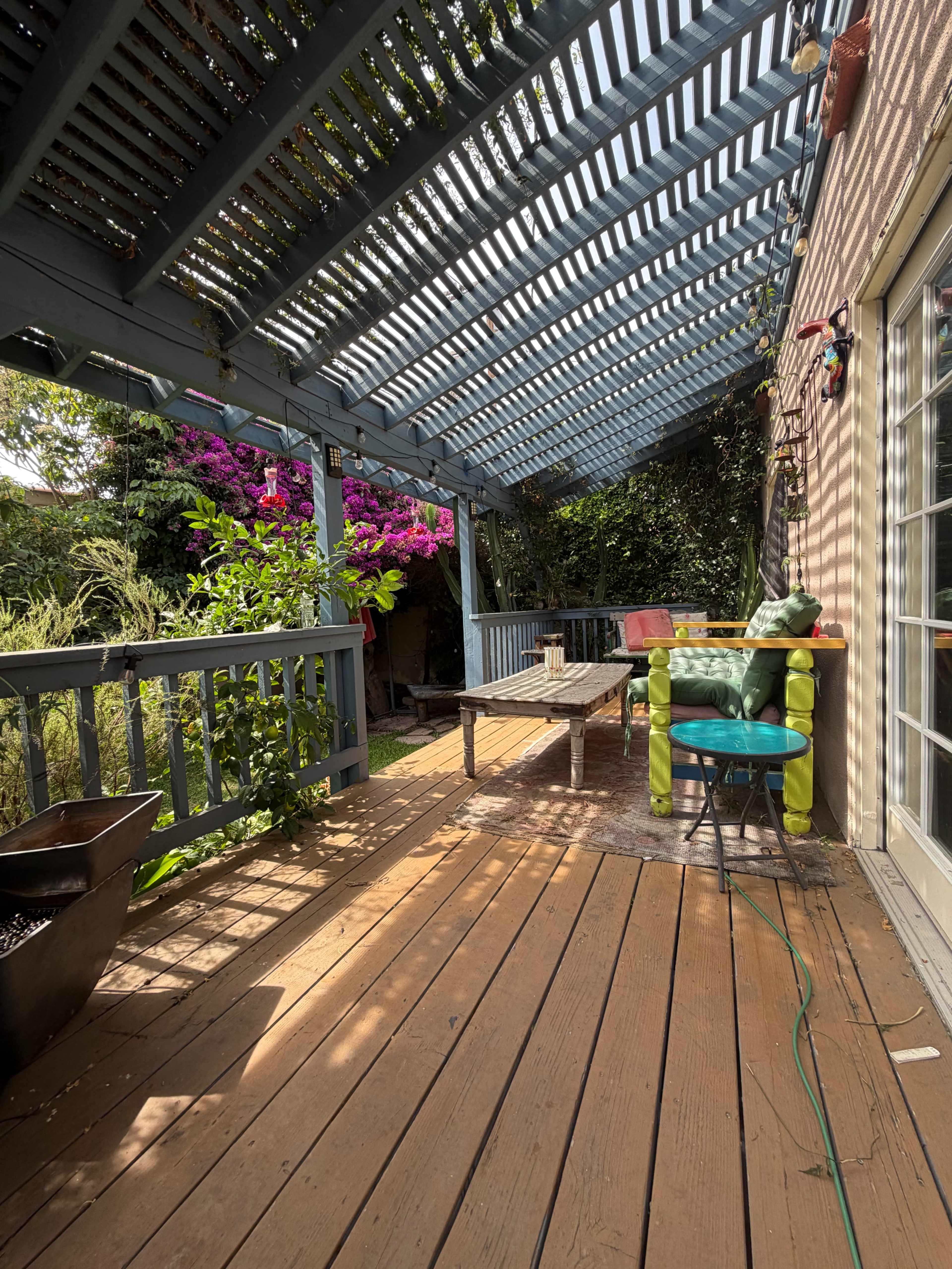The image shows a wooden patio with a trellis overhead, surrounded by greenery and pink bougainvillea, featuring a table and colorful chairs.