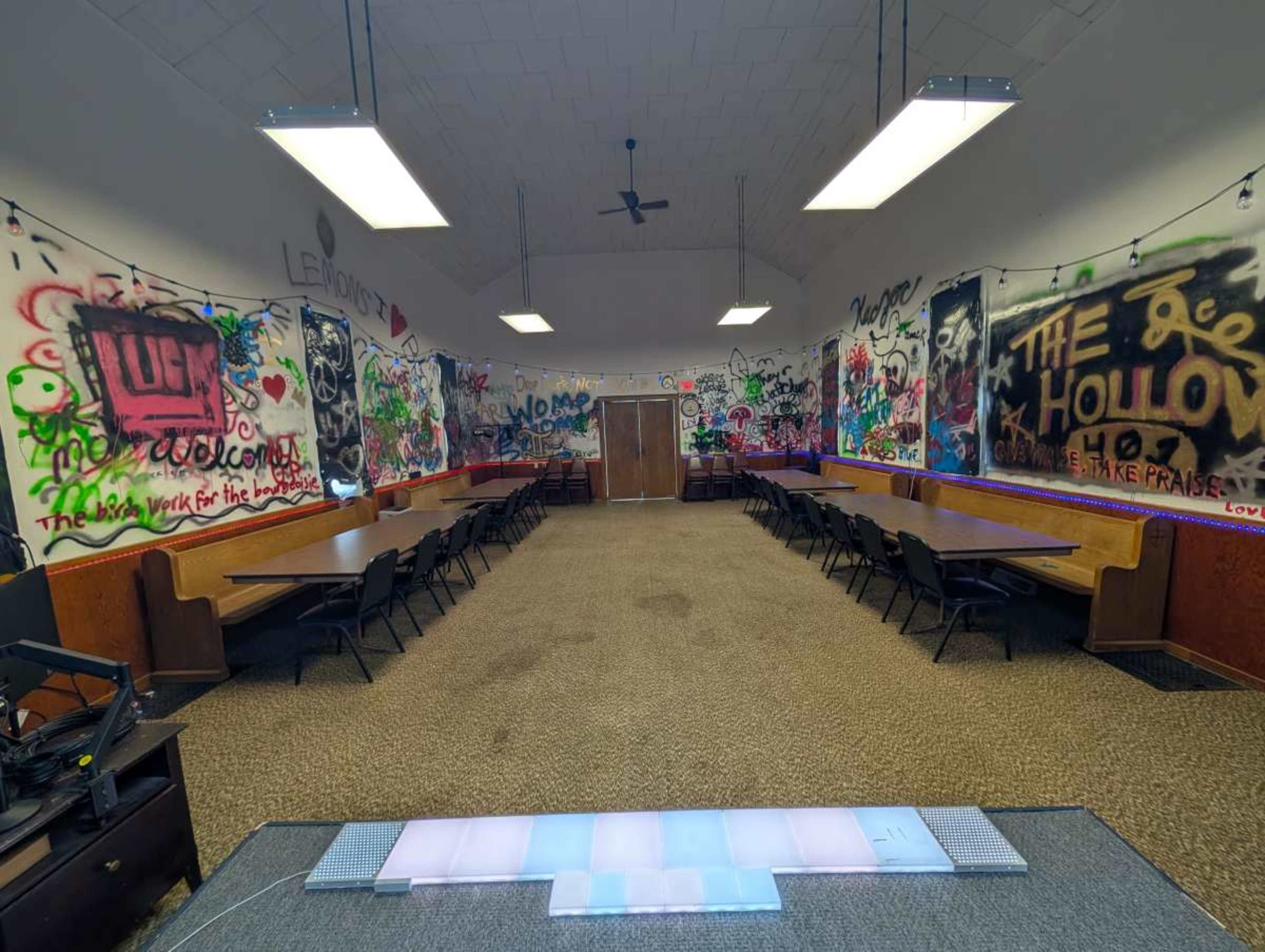 The image shows an indoor space with graffiti-covered walls, rows of wooden tables along the sides, and carpet flooring.