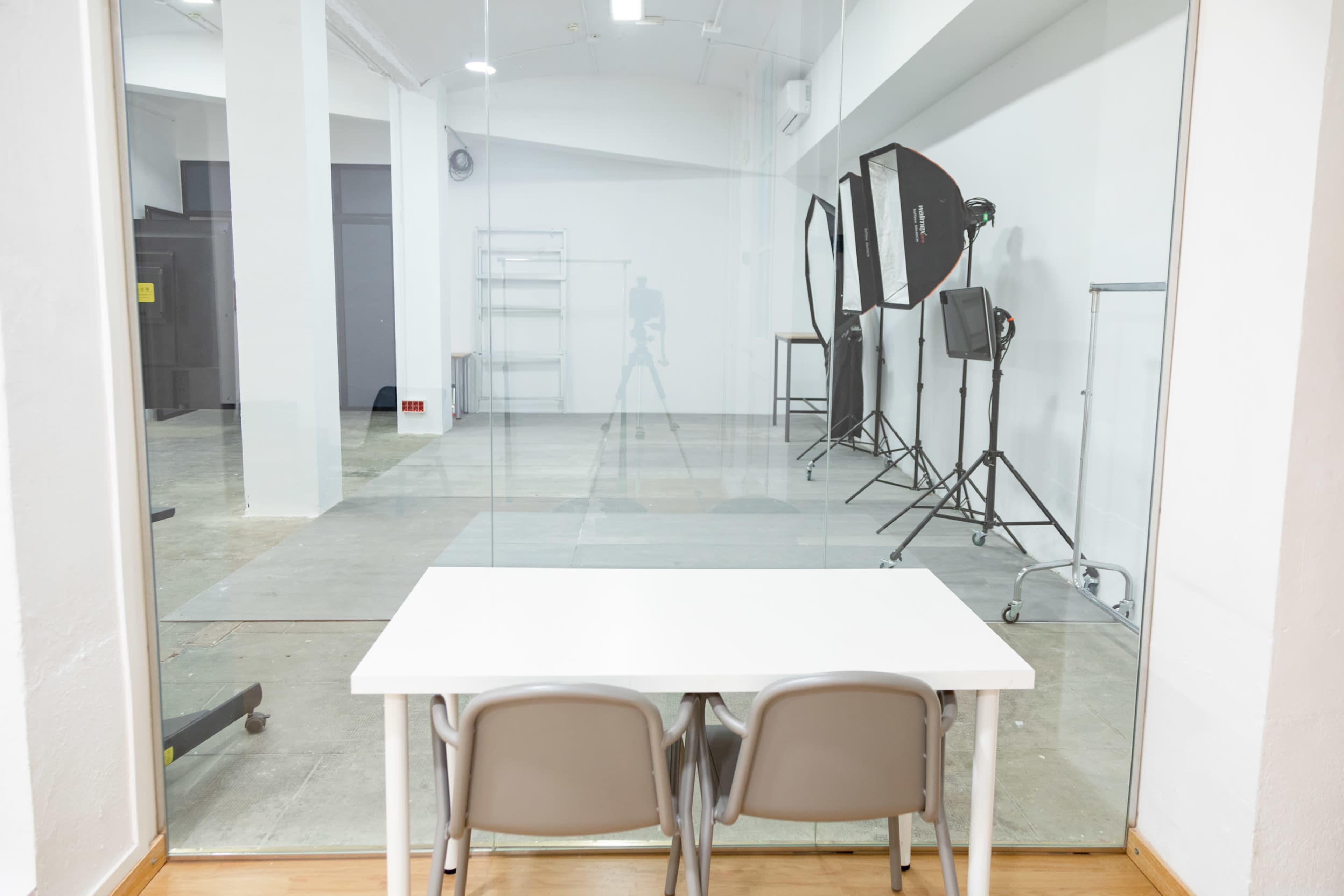 A minimalist studio space is visible through a glass wall, featuring a table with two chairs and photography equipment set up in the background.