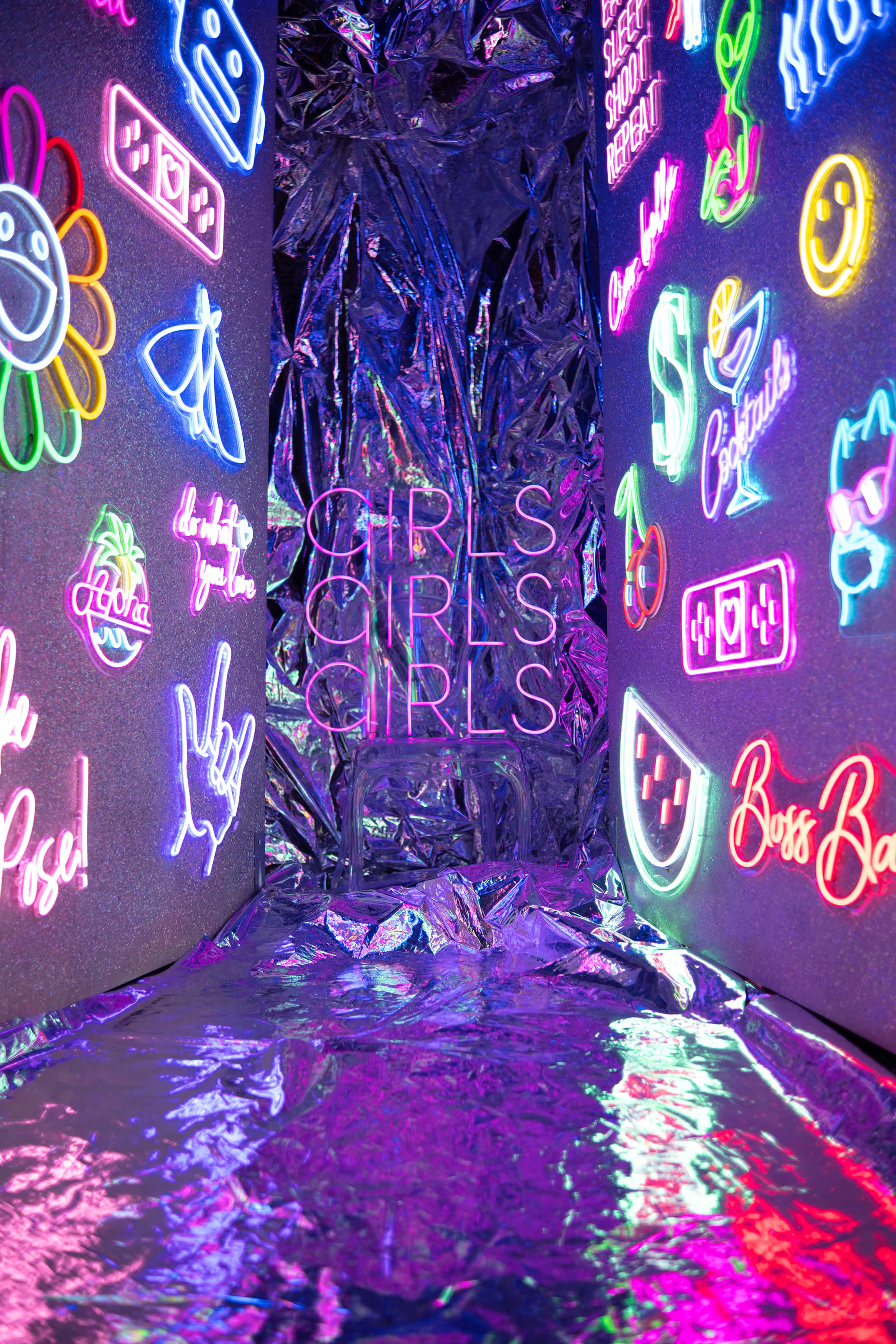 The image features a brightly lit room adorned with various colorful neon signs on the walls, including the phrase "GIRLS GIRLS GIRLS" prominently displayed.