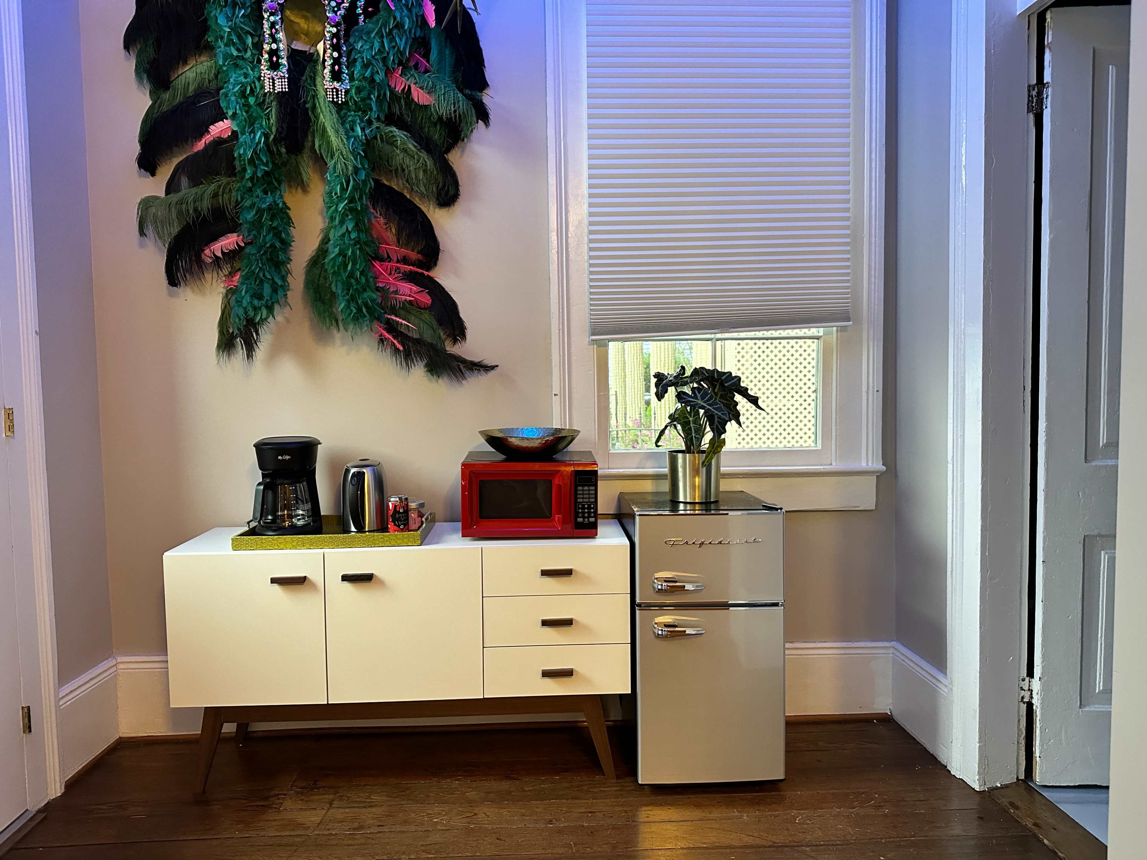 The image shows a kitchen corner with a white cabinet, a coffee maker, a red microwave, a decorative bowl, a small silver refrigerator, and a wall adorned with large, colorful feathers.