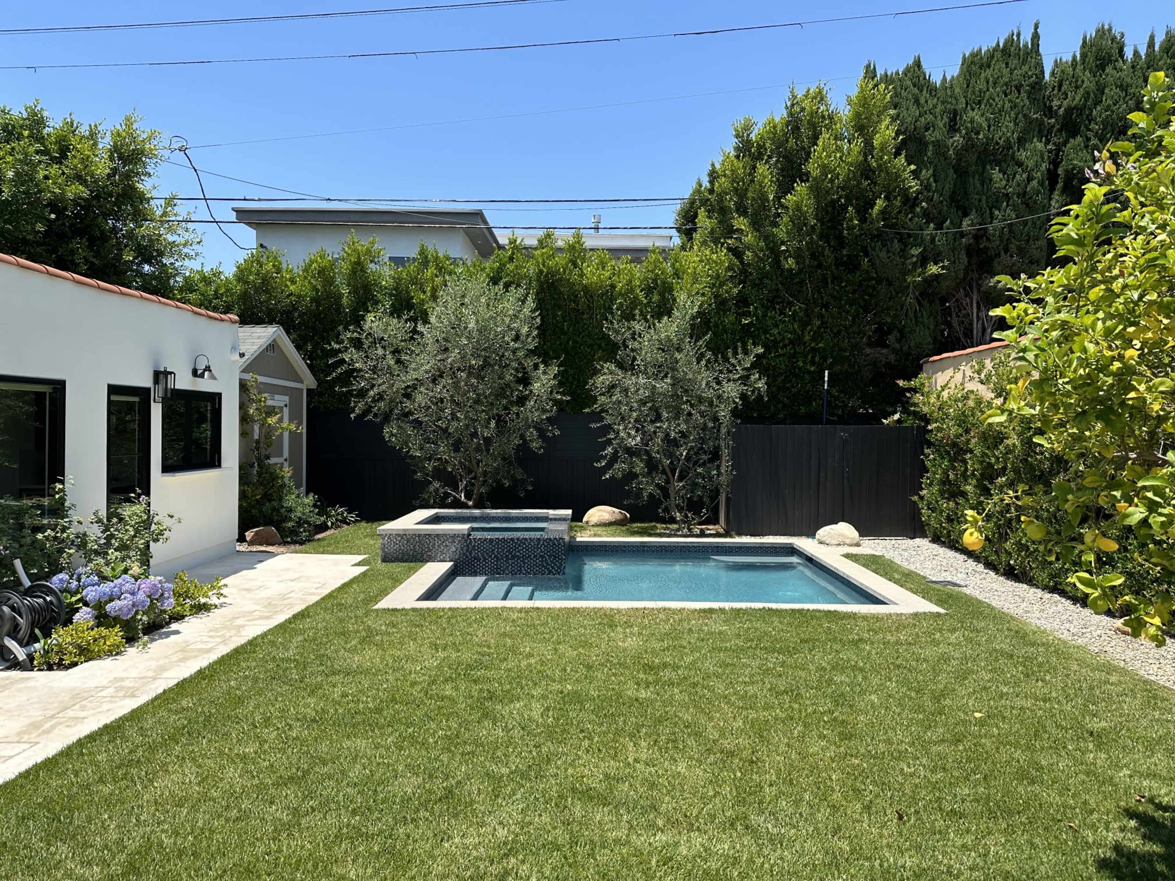 A landscaped backyard featuring a rectangular swimming pool, a small hot tub, and neatly trimmed grass surrounded by trees and shrubs.