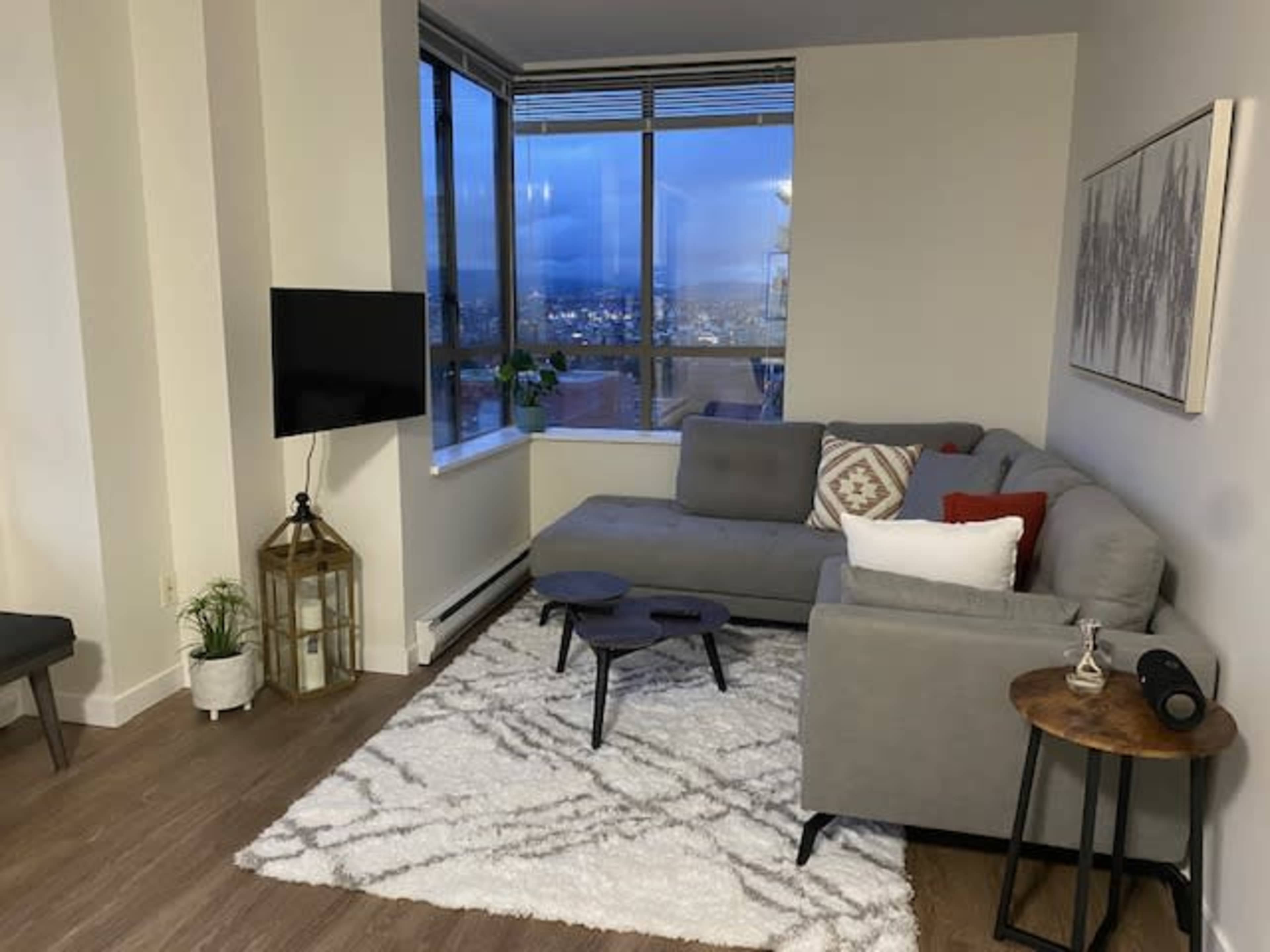 A modern living room features a sectional sofa, a small coffee table, a television mounted on the wall, and large windows with a city view.