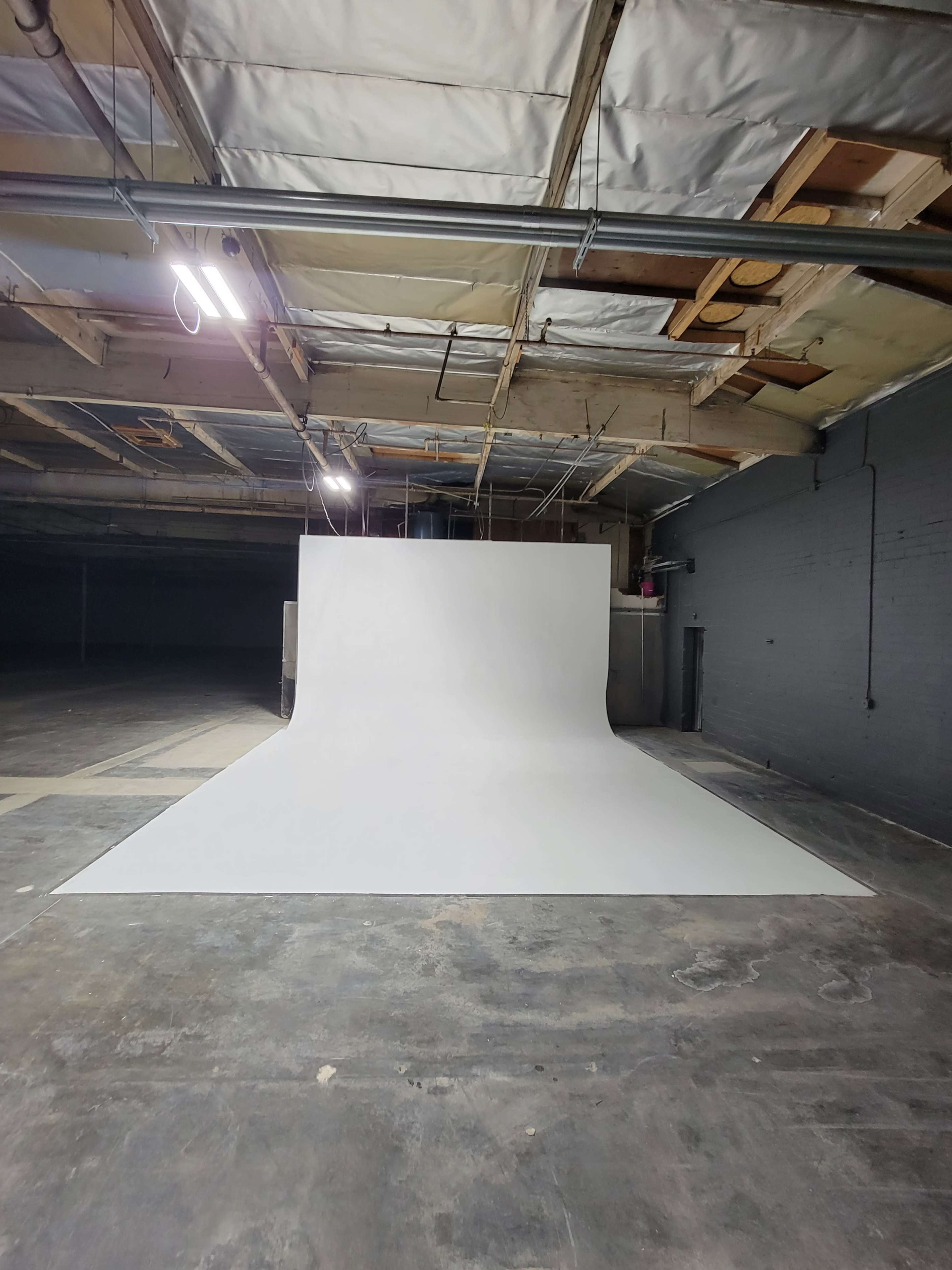 A large, white seamless backdrop is set up in a dimly lit industrial space with exposed beams and concrete flooring.