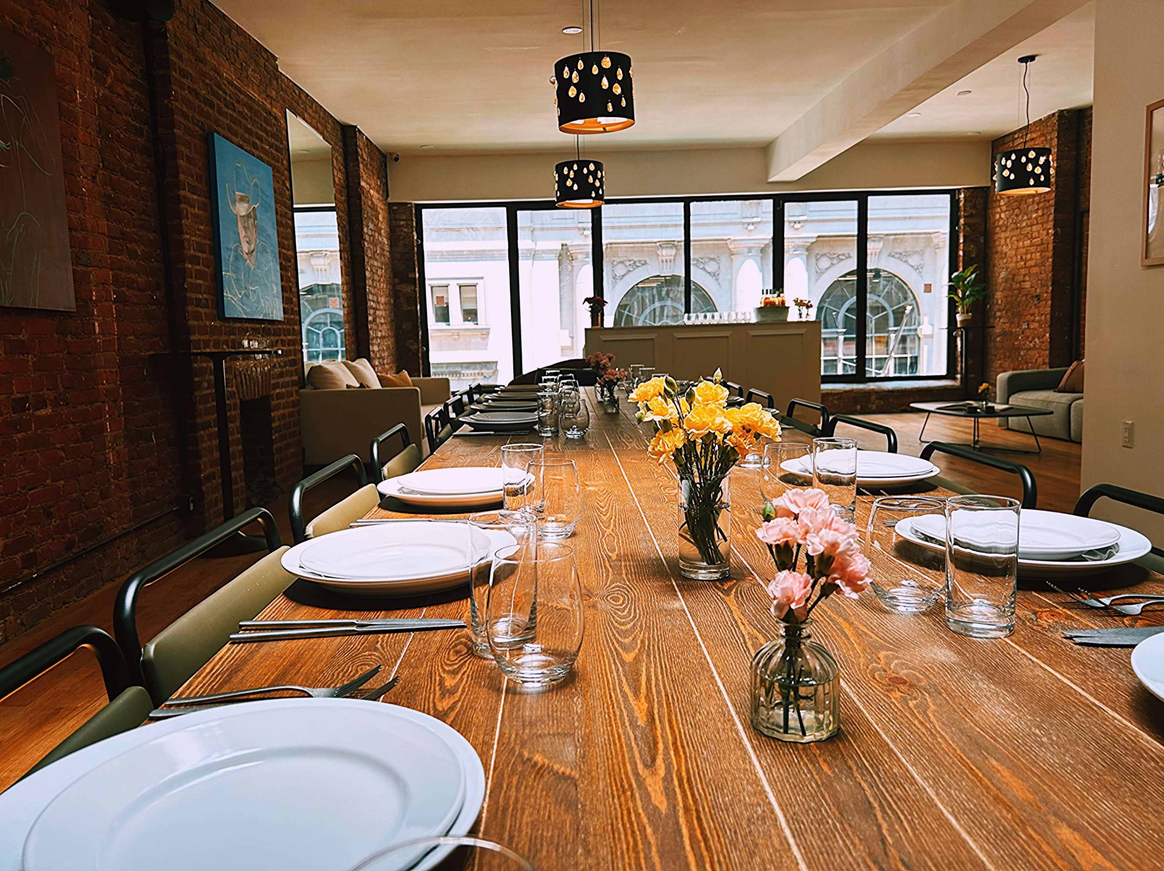 A long wooden dining table is set for a meal with flower arrangements and glassware, situated in a room with large windows overlooking a cityscape.