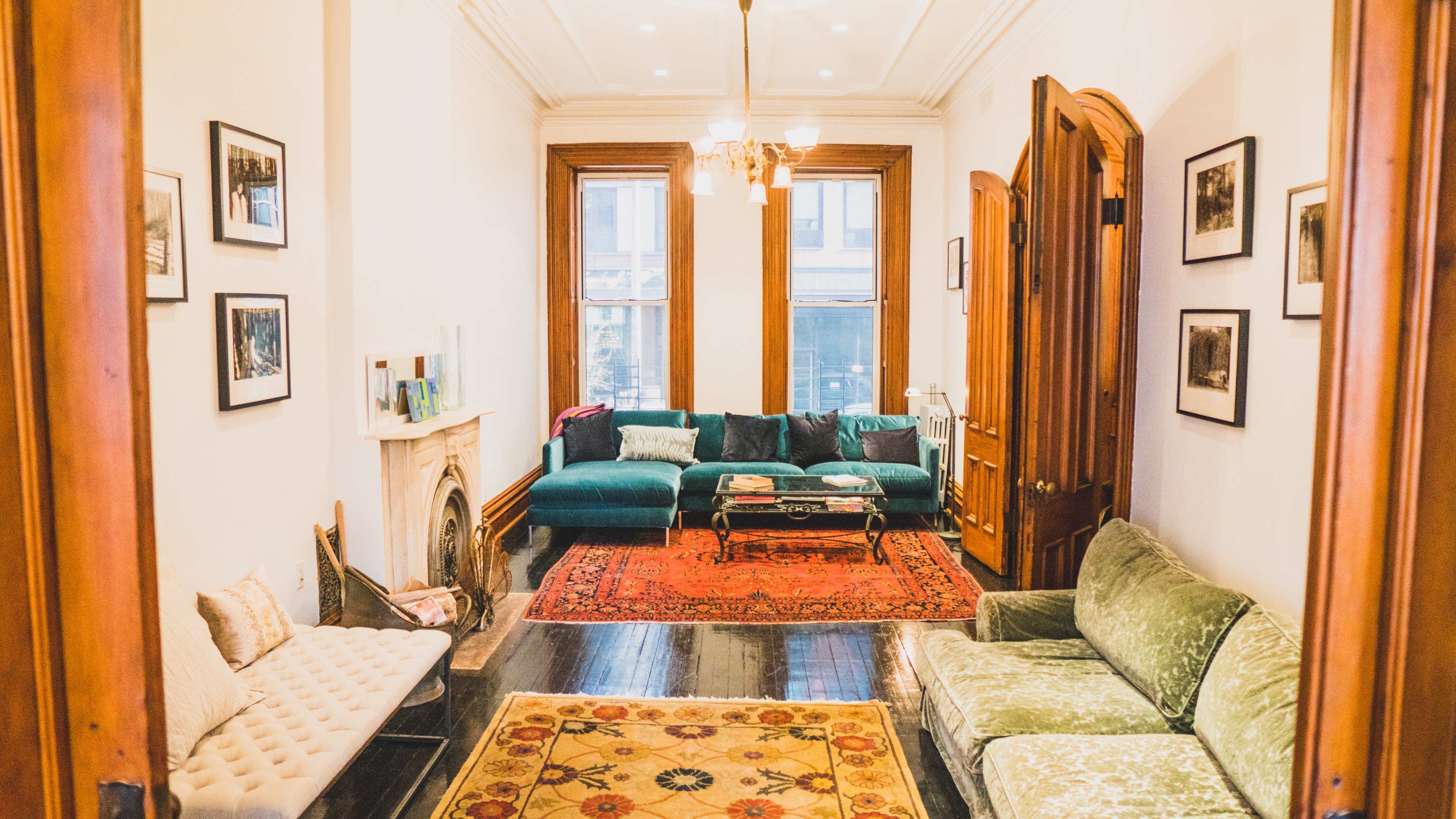 The image shows a bright living room with a mix of green sofas, ornate rugs, and framed photographs on the walls.